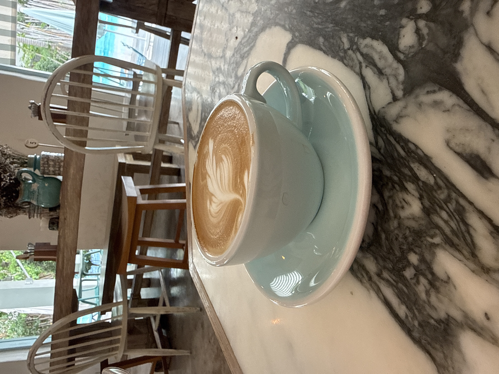 A close-up, eye-level shot features a light blue ceramic cup and saucer with latte art on a marble tabletop. The cup holds a light brown latte with intricate white foam art in the center. The saucer has a light blue hue and a subtle white rim. The tabletop displays a marbled pattern of white and dark grey. In the background, several white wooden chairs with slatted backs are visible around a rustic wooden table, suggesting a cafe or restaurant setting. Through an open doorway or large window, a swimming pool with turquoise water and palm trees are discernible, hinting at an outdoor or resort-like environment. The overall ambiance suggests a tranquil cafe in Hua Hin City Municipality, Thailand.