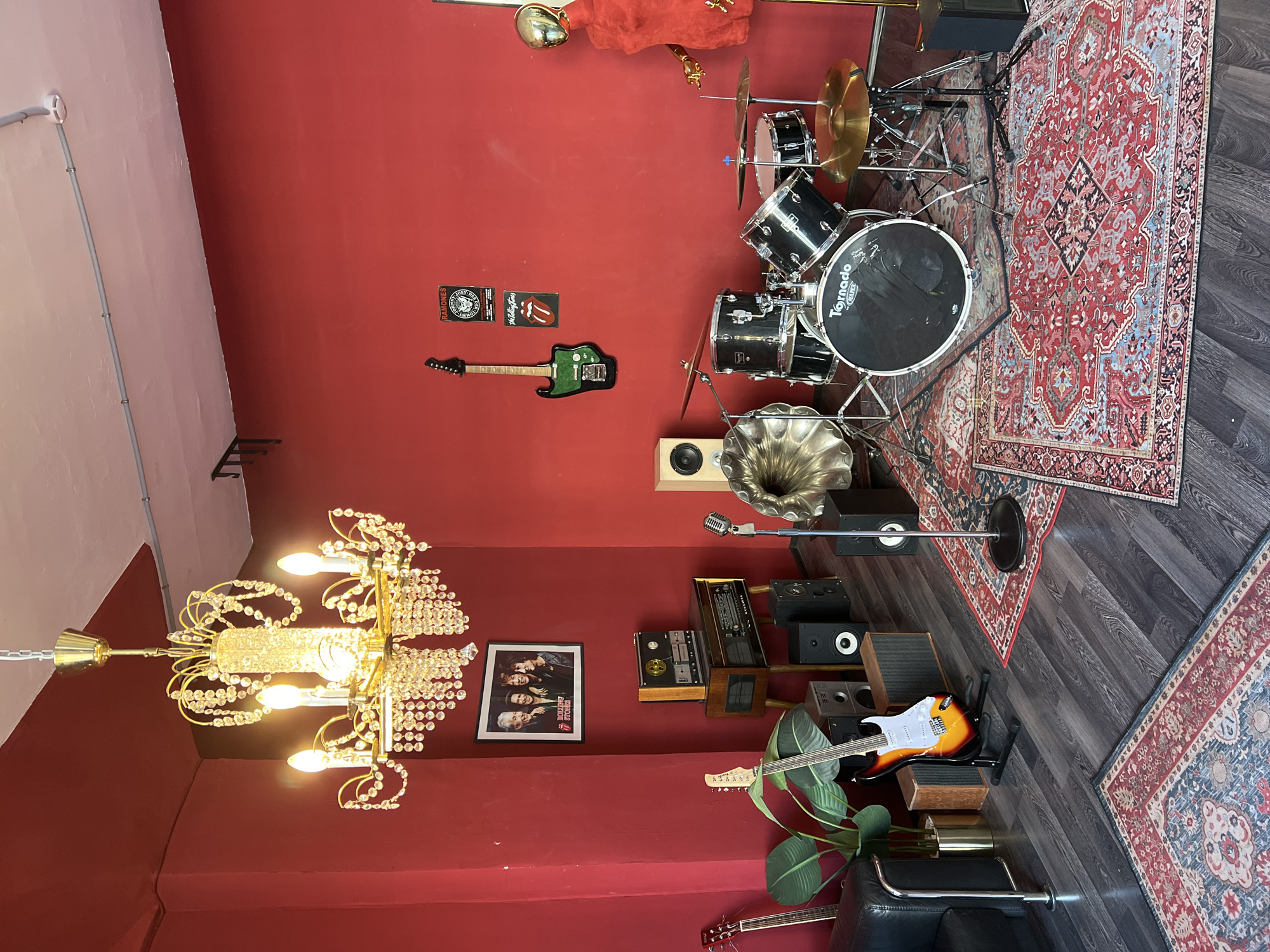 A music room featuring a drum set, guitars, and vintage audio equipment is located in Saint Petersburg, Russia. The room has red walls and a wooden floor covered with Oriental rugs. A large chandelier hangs from the ceiling. There is a framed poster of The Rolling Stones on the wall.