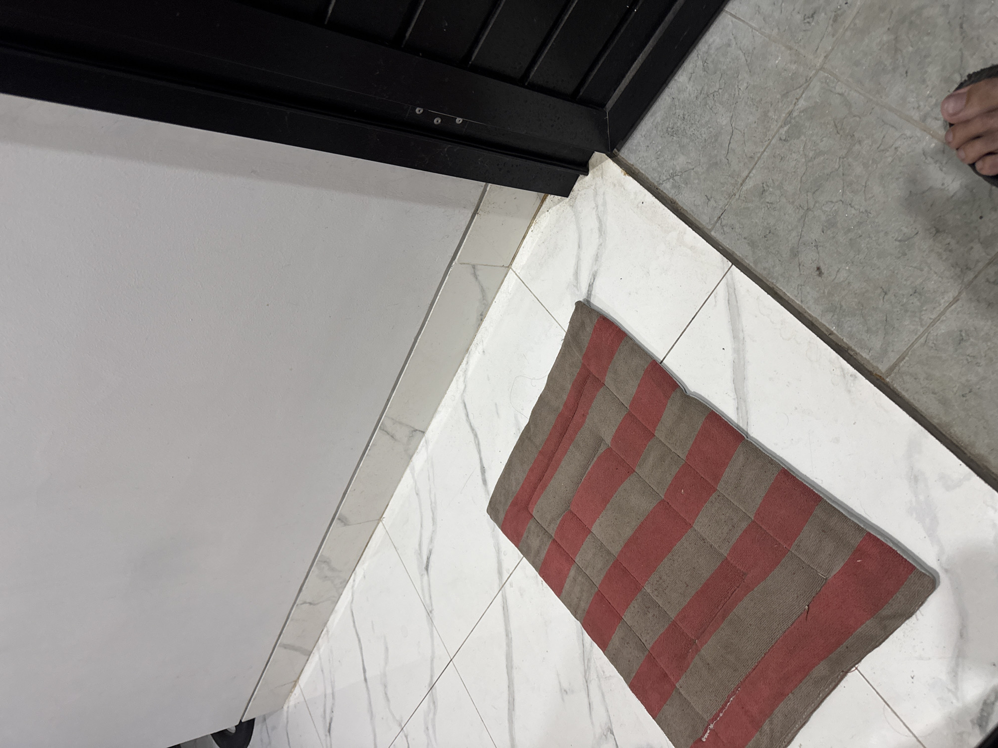 In a room with white marble tile flooring, a red and brown striped rug is placed on the floor. The rug is rectangular and positioned diagonally across the frame. To the right of the rug is a black metal doorframe. The wall is painted white and has a white baseboard. The overall scene appears to be in a residential or commercial building in Sukawati, Indonesia, a location known for its cultural heritage and crafts.