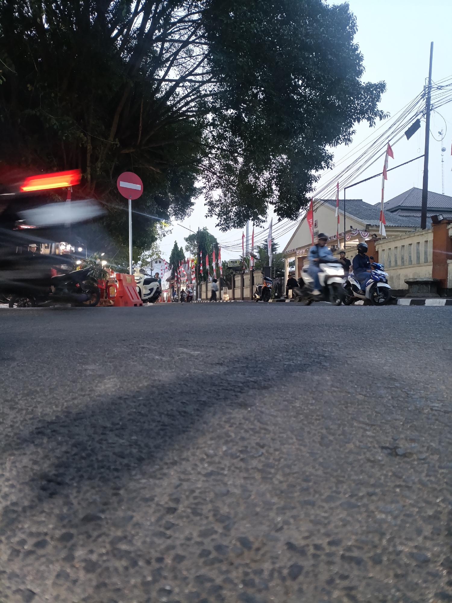 The image captures a street scene in Jonggol, Indonesia, during what appears to be late afternoon or early evening. The perspective is low, taken from the surface of an asphalt road.
In the foreground, the road's textured surface is prominent. A dark shadow, likely cast by a large tree to the left, stretches across the asphalt.
To the left, a motorcycle, partially obscured and blurred due to motion, is visible. Its red taillight is streaked horizontally across the frame, indicating movement. Beside it, a red and white traffic barrier is placed on the road. A red circular traffic sign with a white horizontal bar is mounted on a pole behind the barrier. A large tree with dense foliage dominates the upper left quadrant of the image, its branches extending towards the sky.
Further down the street, several people are on motorcycles, also showing signs of motion blur. One white motorcycle with a rider is moving towards the right. To its right, a blue and white motorcycle with two riders is also in motion.
The background features a building with a light-colored facade and a tiled roof, decorated with Indonesian flags and pennants. A concrete wall runs along the side of the building, with stone pillars at intervals. Utility poles with numerous wires are visible against the sky, which is overcast and a pale gray. The overall impression is of a moderately busy street in a town or city, possibly a public area given the flags and building architecture.