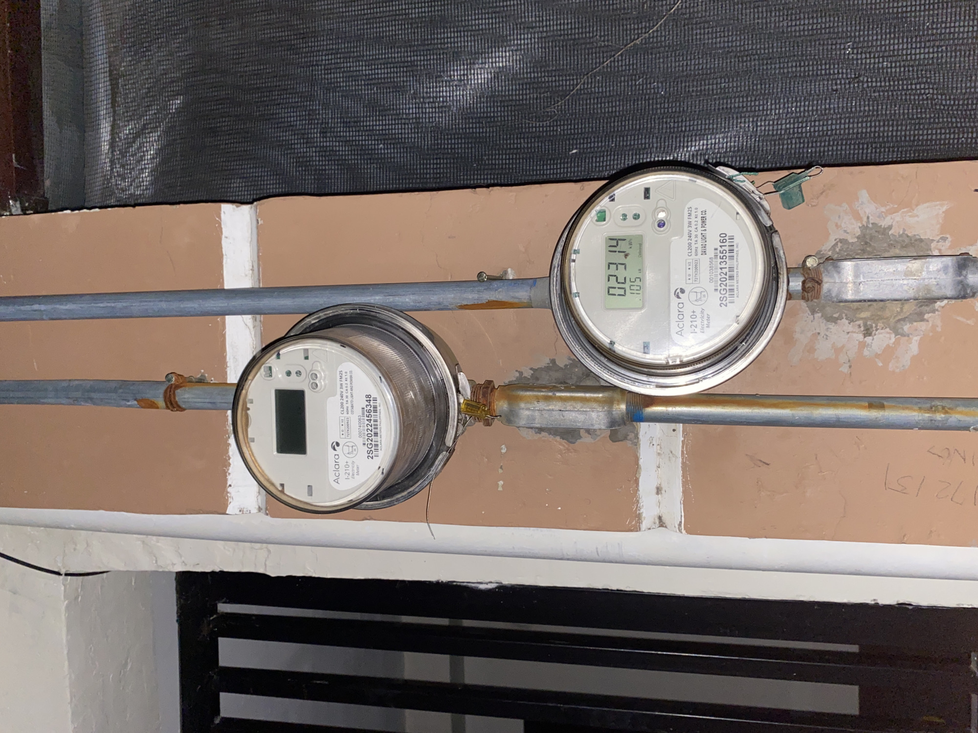 The image shows two Aclara electricity meters mounted on a wall in Davao City, Philippines. The meters are vertically aligned and attached to a metal conduit. The upper meter has a blank digital display. The lower meter displays "023.14 kWh" and "105 kW". The wall is a light brown color with some visible plaster and discoloration. To the left of the meters is a dark-colored metal grate. To the right is a dark-colored mesh material. There is also some graffiti or writing on the wall to the left of the lower meter. The scene appears to be outdoors, likely on the exterior of a building.