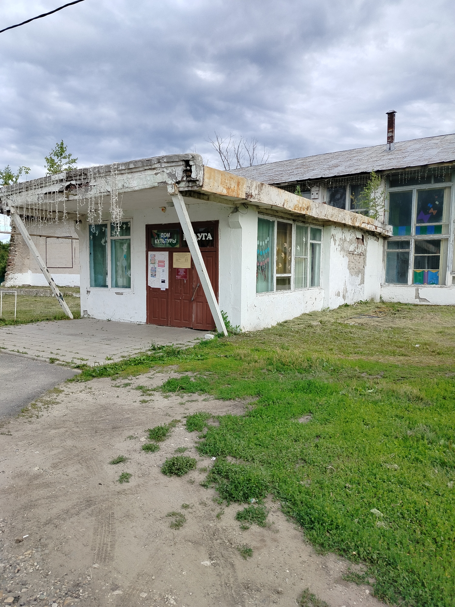 The image shows a small, weathered building, likely a community center, in the village of Grabovo, Russia. The building is painted white and has a rusty, partially collapsed overhang adorned with what appear to be old Christmas lights. A brown door with two windows is visible, with posters displayed on it and the wall next to it. The building has several windows, some with decorative elements visible inside. The structure appears to be in a state of disrepair, with peeling paint and crumbling concrete. The surrounding environment is grassy, with patches of dirt and tire tracks indicating vehicular access. The sky is overcast. The building's facade has Cyrillic text that reads "ДОМ КУЛЬТУРЫ" which translates to "House of Culture."