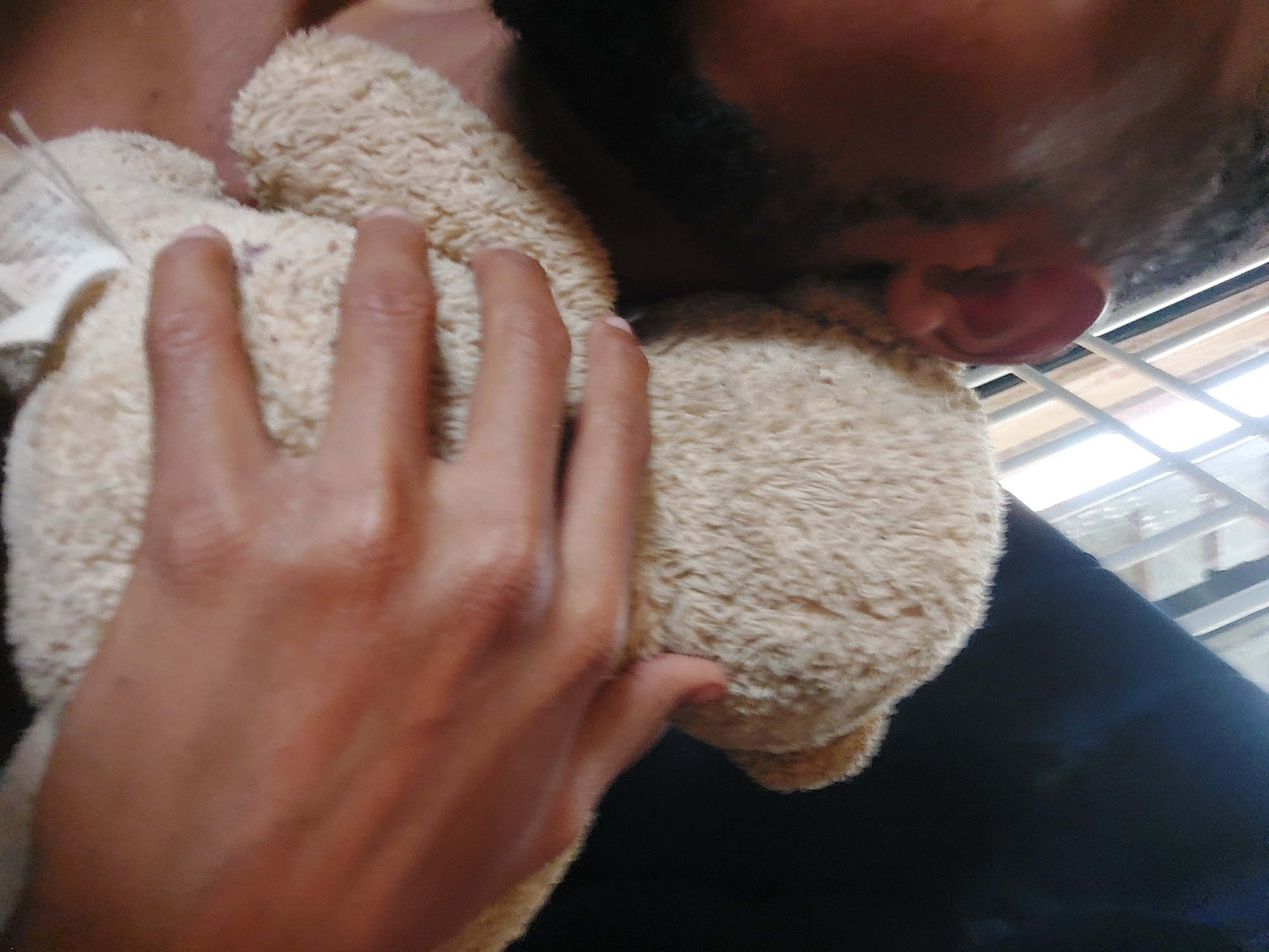 A person is holding a tan teddy bear to their face. The person has dark hair and a beard. Their ear is visible. The person's hand is on the teddy bear. Tags are visible on the bear. The background is blurred. The location cannot be determined from the image.