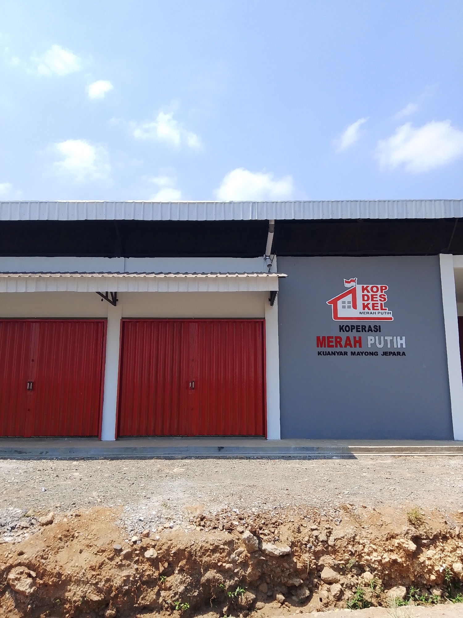 The image shows a building with two red rolling shutter doors on the left and a grey wall with a logo and text on the right. The building has a white corrugated metal roof and a dark grey overhang. The logo on the grey wall is a stylized house with a flag and the text "KOP DES KEL MERAH PUTIH". Below the logo, it says "KOPERASI MERAH PUTIH" in large red letters, and "KUANYAR MAYONG JEPARA" in smaller black letters. The area in front of the building is a dirt and gravel lot with some construction visible in the foreground. The sky above is blue with scattered white clouds. The location is identified as Kuanyar, Mayong, Jepara, Indonesia.