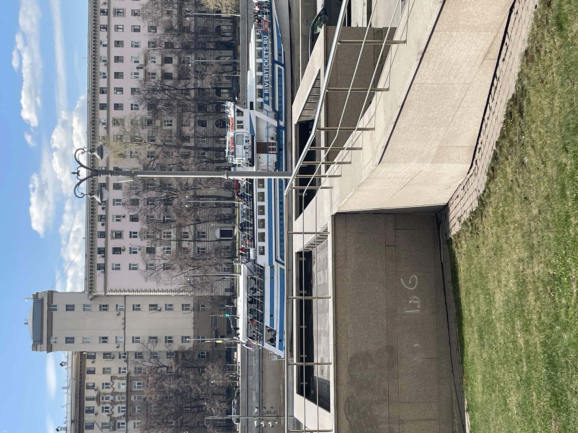 The image depicts a street scene in Moscow, Russia, angled vertically. On the left side, there are several tall, multi-story buildings in a pinkish hue. Streetlights are visible in front of the buildings. A riverboat, with the name "RIVERTICKETS.RU" on its side, is docked. People are on board. The right side of the image includes a modern architectural structure with metal railings, a concrete walkway, and a patch of green grass. Graffiti is on the concrete wall. The sky is blue with scattered clouds. Trees are bare.