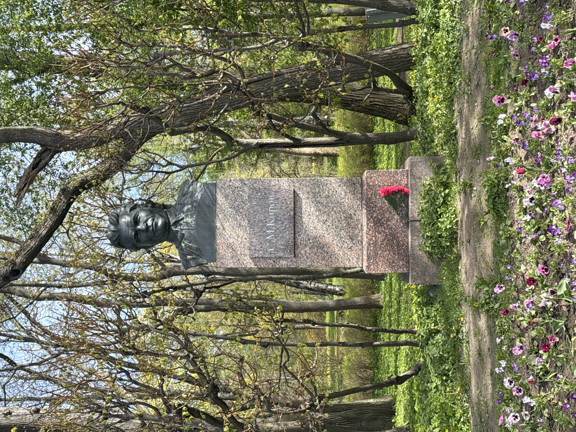 The image depicts a bust of Sergei Kirov, located in Saint Petersburg, Russia. The bronze bust sits atop a granite plinth inscribed with "С.М. Киров" (S.M. Kirov). Red carnations lie at the base of the monument. The bust is surrounded by trees with budding leaves and a flower bed containing pansies. A worn dirt path divides the flower bed.