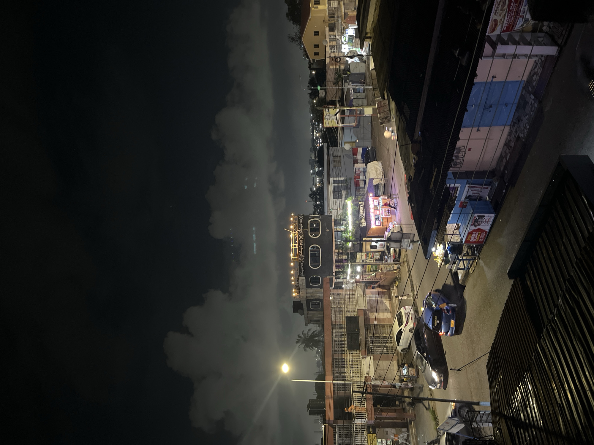 The image depicts a nighttime street scene in Accra, Ghana. The sky is dark with intermittent clouds, illuminated by artificial lights from streetlights and buildings. In the foreground, a corrugated metal roof of a building is visible. The street below is paved and wet, reflecting the light.
Several vehicles are present on the street. A dark colored sedan is parked on the left, and a bright blue hatchback is parked next to it. Further down the street, a white sedan is parked, and a dark colored SUV is also visible. Pedestrians are scattered throughout the scene. One person in an orange garment stands on a balcony of a building on the left. Other figures are walking on the sidewalk or near the parked cars.
Buildings of varying heights and architectural styles line the street. On the left, a multi-story building features balconies and a prominent streetlight. To the right of this, a building with an unusual, rounded facade is illuminated with string lights along its roofline and displays the text "Johnny's Pub". Further to the right, a yellow, two-story residential building with a red roof is visible. In the middle ground, smaller shops and stalls are illuminated by commercial lighting, some with awnings. The overall impression is a lively urban environment at night, with signs of commercial activity and residential life.