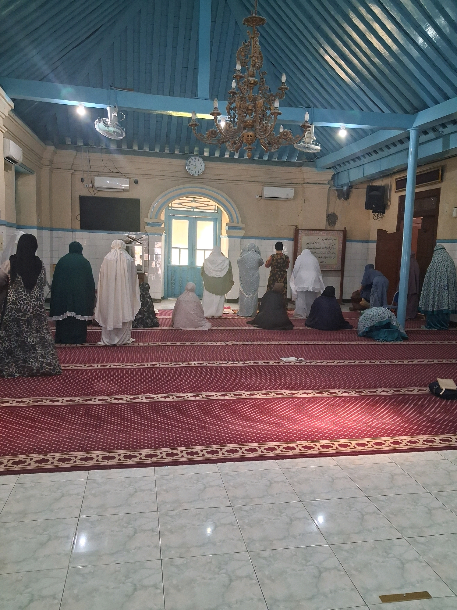 The image depicts the interior of a mosque in Surakarta, Indonesia, likely the Kauman Great Mosque due to its architectural style and historical significance. The mosque features a blue-painted wooden ceiling with exposed beams and a large, ornate chandelier. The walls are light-colored, and a clock is visible on the far wall. A patterned red carpet covers the floor, with a white tiled area in the foreground. Several women are present, dressed in Islamic attire, engaged in prayer. Some are standing, while others are kneeling or prostrating on the carpet. The women are facing the qibla, which would be towards Mecca. The overall atmosphere appears serene and devotional.
