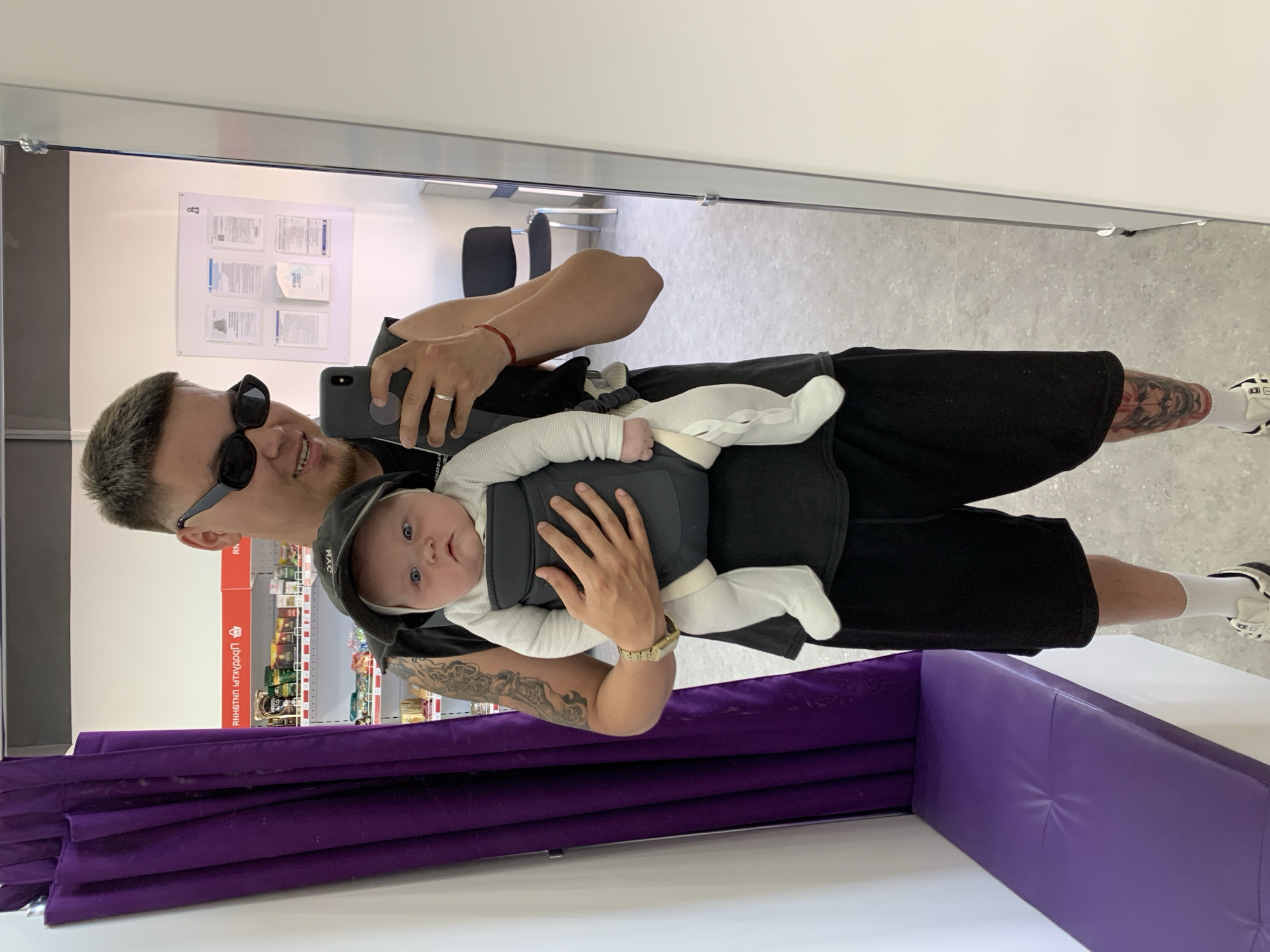In a retail environment, likely a fitting room or a changing area within a store, a man is seen taking a mirror selfie while holding a baby in a carrier. The man is wearing sunglasses, a black t-shirt, and black shorts. He has tattoos on his arm and leg. The baby is wearing a white outfit and a black hat. In the background, shelves stocked with various products are visible, suggesting a retail setting. A purple curtain hangs to the left of the frame, and a purple bench is present in the foreground. The location is identified as a store in Stavropol, Russia, likely a clothing or general merchandise store given the fitting room and product displays.