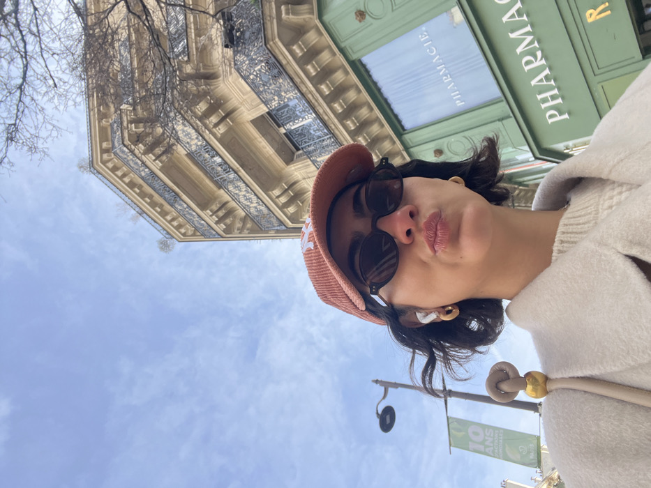 A low-angle selfie captured on a city street in Paris, France, features a female subject positioned slightly off-center to the left. The subject, seen from the chest up, wears a rust-colored corduroy baseball cap with a white "NY" logo, dark oval sunglasses, white wireless earbuds in both ears, and small gold-colored hoop earrings. Her dark, wavy hair is visible, and her lips are puckered in a kiss gesture. She is dressed in a light-colored, possibly cream, ribbed knit sweater and a matching overcoat, with a light-colored shoulder bag featuring a unique knot and gold sphere detail visible on her left shoulder.
The background reveals a clear blue sky with sparse clouds. To the right, a multi-story Haussmannian-style building with a light-colored facade and ornate dark metal balcony railings occupies the upper frame. Below this building, a distinct green storefront with the word "PHARMACIE" in prominent white capital letters indicates a pharmacy at street level. On the left side of the frame, a black lamppost is visible, and below it, a green banner or flag attached to a pole displays "10 ANS" and partially visible "PARIS" text.