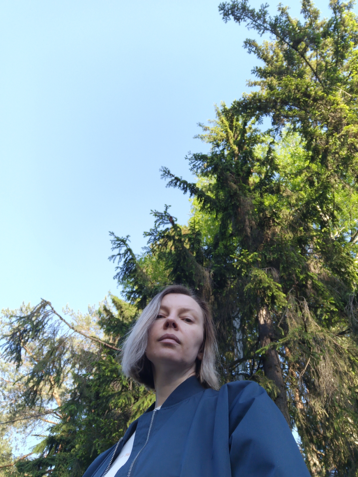 A low-angle, medium shot shows a woman standing in front of a cluster of trees in Chaikovsky, Russia. A clear, blue sky is visible in the upper left. The woman has fair skin, short light-brown/blonde hair, and is wearing a dark blue zip-up jacket over a white shirt. The trees behind her appear to be pine trees with long, drooping needles. The location is Chaikovsky, Russia.