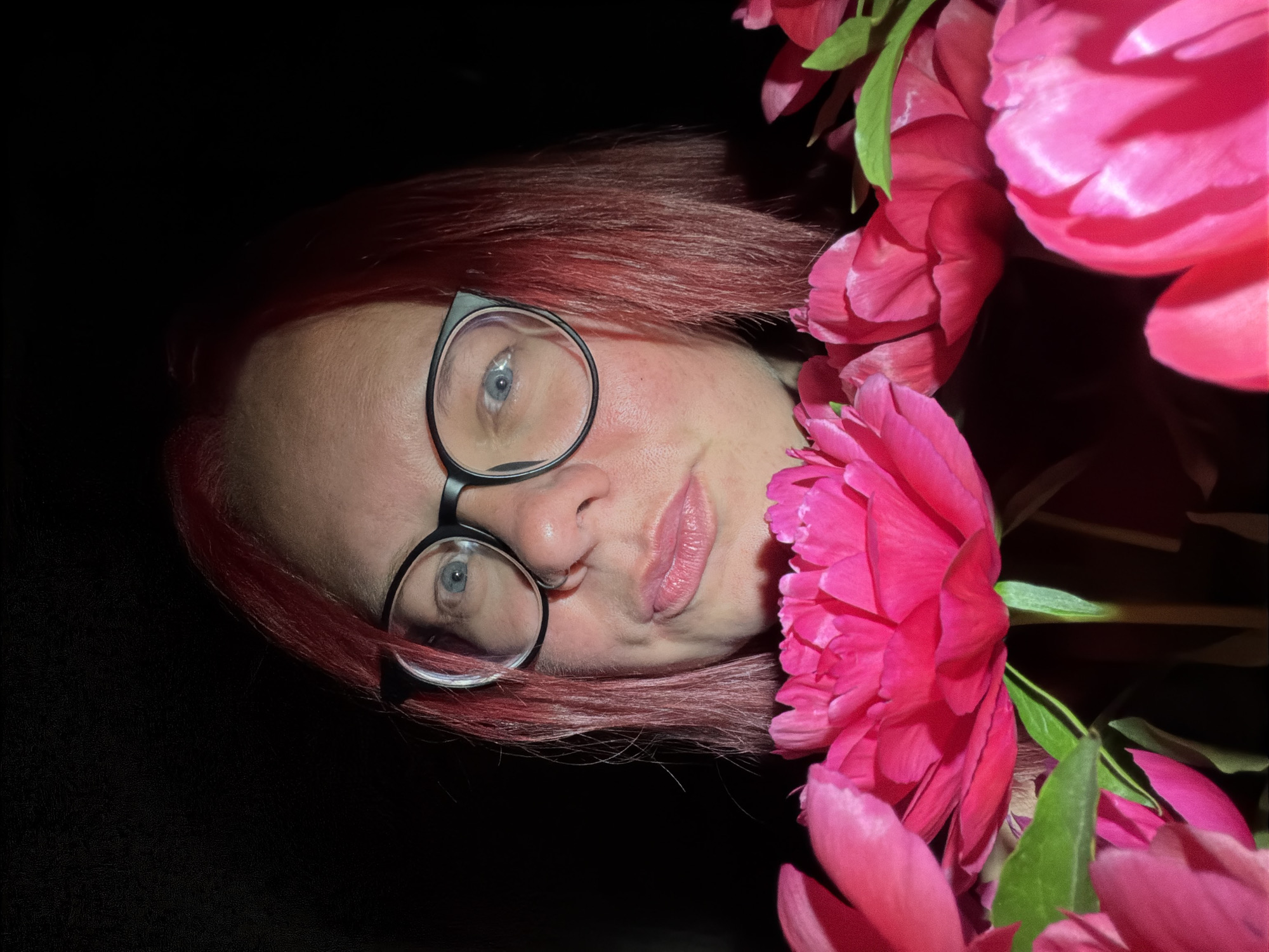 A close-up, low-light portrait captures a person, likely female, with shoulder-length, dark pink/red hair. They wear black-framed eyeglasses, through which light blue eyes are visible. The individual has a fair complexion with reddish undertones on the cheeks and forehead. Their lips are closed, forming a slight, upward curve on the right side, suggesting a subtle smirk or contemplative expression. The person's head and upper chest are positioned in the upper half of the frame. In the lower half of the image, vibrant, hot pink peonies with multiple layers of petals are prominently displayed, partially obscuring the lower face and body of the individual. Green stems and leaves are visible among the flowers. The background is uniformly dark, indicating either very low ambient light or a deliberately black backdrop.