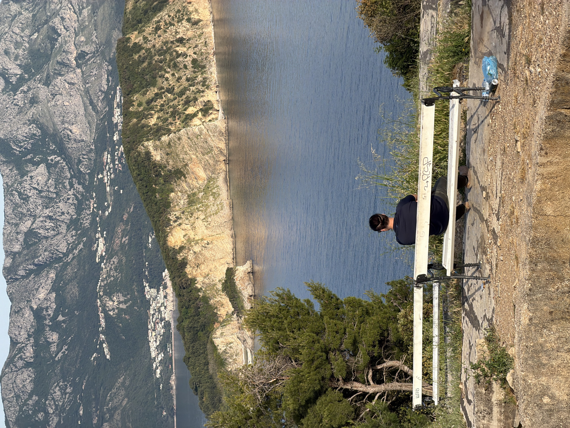 A medium shot shows a man sitting on a white park bench, overlooking a scenic view. The bench is located on a stone ledge with some greenery growing around it. The man is facing away from the camera, and a water bottle in a blue plastic bag sits near the edge. Below the ledge is a body of water surrounded by mountains and cliffs, some of which are covered in vegetation. A small settlement of buildings is visible on the mountainside. The image is taken during the day, with ample sunlight. The location may be in or near Budva, Montenegro, based on the terrain and coastal features. The image has been rotated 90 degrees counterclockwise.
