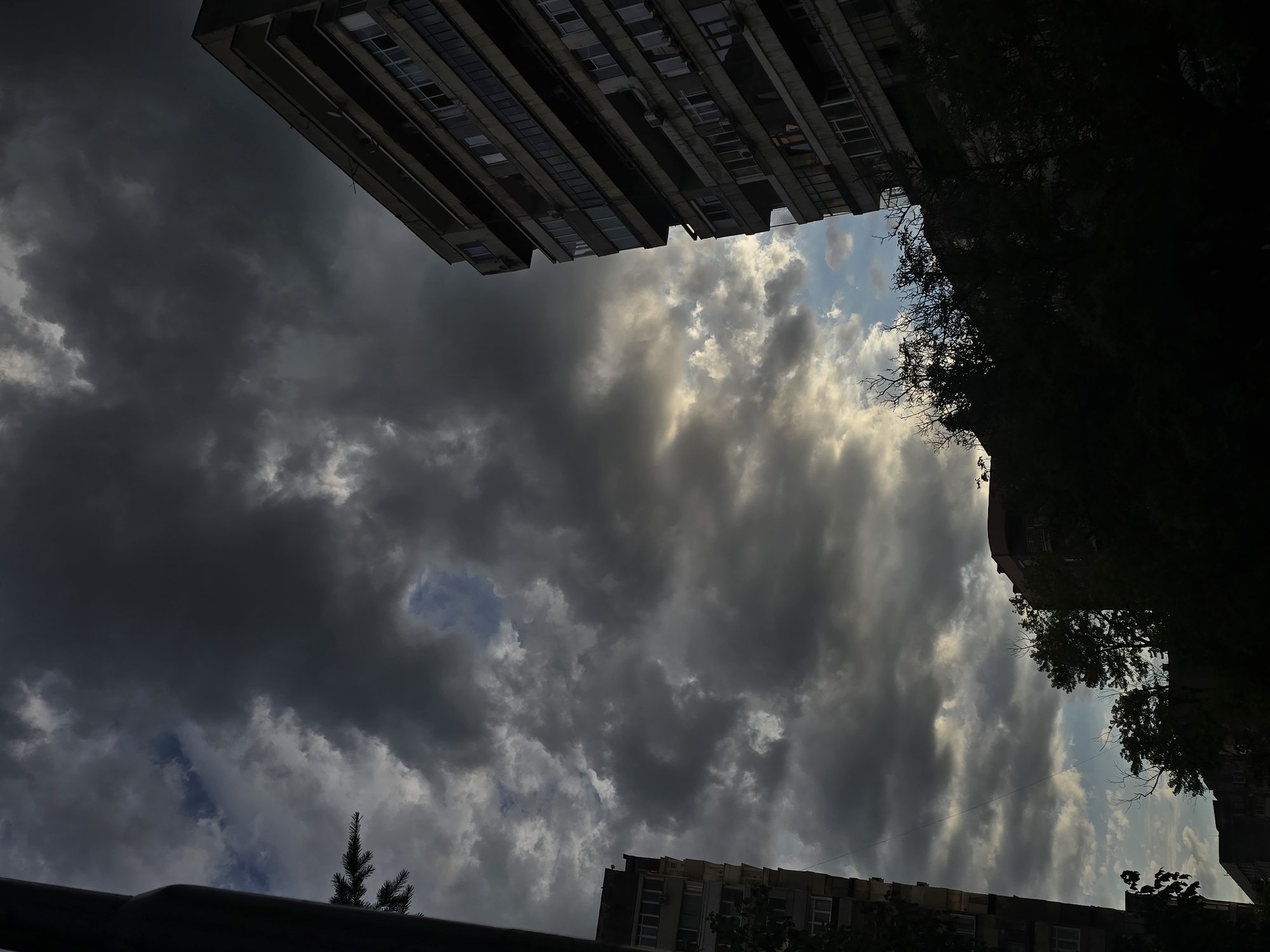 The photograph captures a view of buildings and the sky in Ruse, Bulgaria, under a dramatic, overcast sky. The lighting suggests it is either early morning or late evening, with the sun partially breaking through the clouds, creating illuminated patches.
On the right side of the frame, a tall, brutalist-style apartment building dominates the view. It is made of concrete and has numerous balconies and windows, many of which appear dark or unoccupied. The building's facade is weathered, showing signs of age.
To the left, another apartment building is partially visible, also appearing to be concrete construction with multiple floors and windows. A dark vertical element, possibly a utility pole or part of another structure, is in the far left foreground, obscuring a small portion of the left building and a branch of a conifer tree.
In the foreground and middle ground, dense silhouettes of trees and foliage obscure the lower parts of the buildings and suggest a leafy urban environment. The trees are dark and appear lush, contrasting with the stark architecture.
The sky is filled with heavy, dark clouds, with breaks of lighter grey and patches of pale blue. The interplay of light and shadow across the clouds adds to the dramatic atmosphere. No people are visible in the image. The scene is static and conveys a sense of urban quietude under a moody sky.