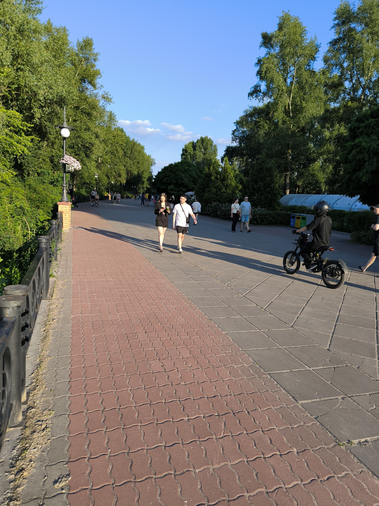 In an outdoor park setting with lush green trees and a clear blue sky, a paved walkway is observed. The walkway is divided into two sections: one with red interlocking pavers and another with grey square pavers. On the grey section, a person wearing a black outfit and helmet is riding a black electric motorcycle, heading away from the camera. Further down the walkway, a couple walks hand-in-hand, with the woman in a black outfit and the man in a white t-shirt and black shorts. Other pedestrians are scattered in the background. The scene appears to be a public park or promenade on a sunny day, with the sunlight casting long shadows across the path.