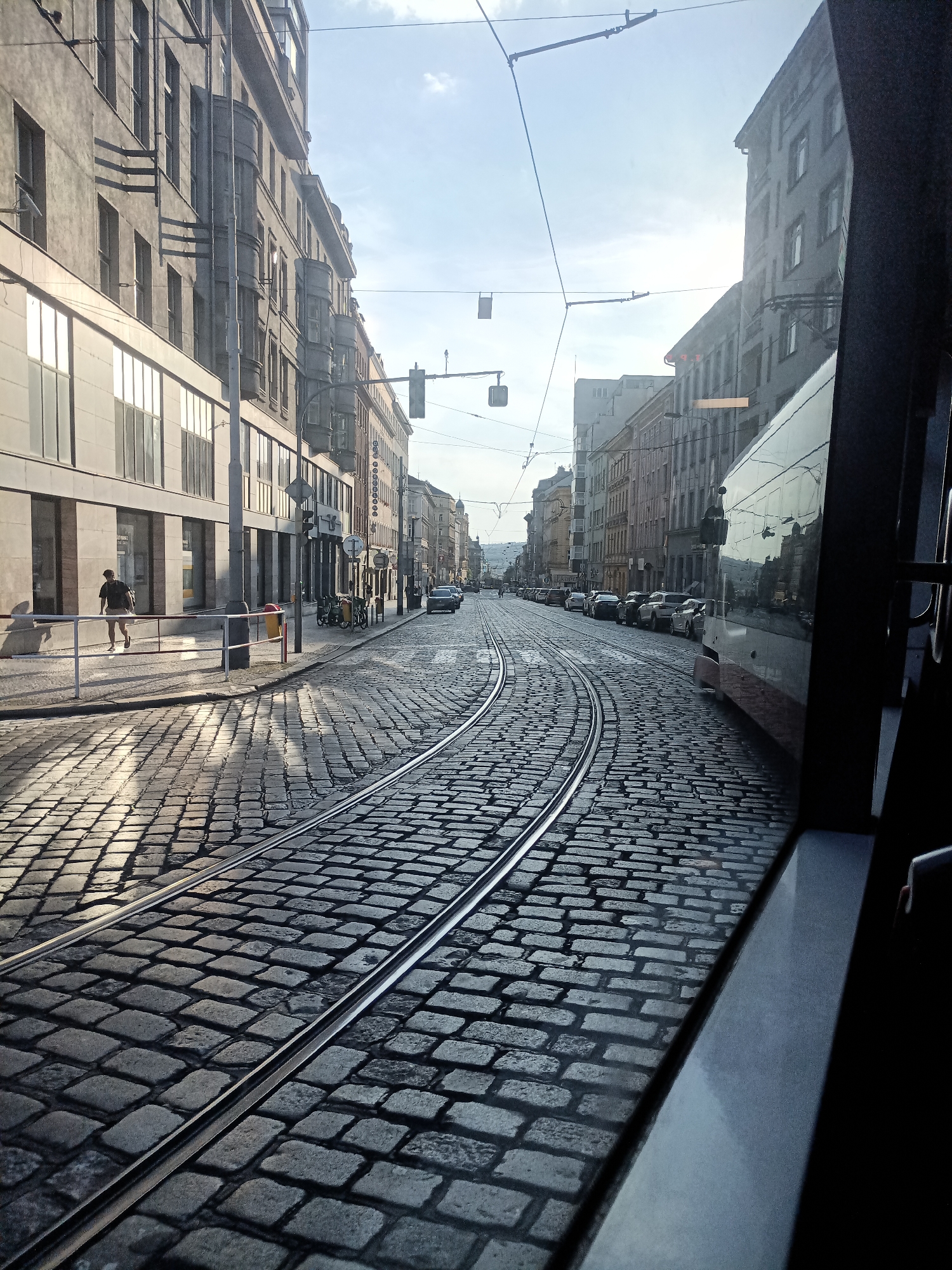 The image captures a street scene in Prague, Czechia, from the perspective of a tram. The street is paved with cobblestones, and tram tracks run down the center, curving to the right. On the left side of the street, there are multi-story buildings with windows and architectural details, and a sidewalk with a person walking. On the right side, the tram's window and interior are visible, reflecting the buildings and sky. Further down the street, more buildings, parked cars, and a traffic light are visible. The sky is bright with some clouds, and sunlight casts shadows on the cobblestones.