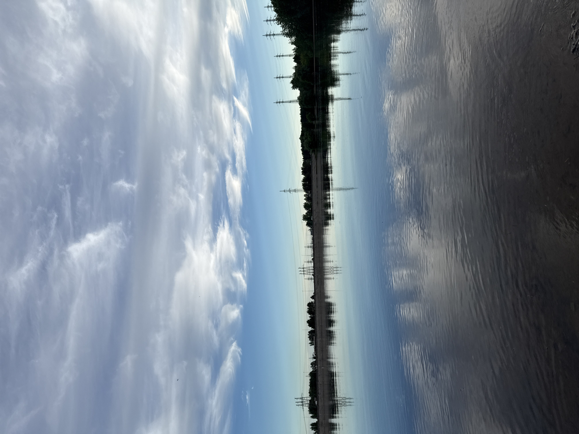 The image depicts a landscape featuring a body of water reflecting the sky, trees, and power lines. Wispy clouds are visible in the sky. The trees and power lines form a horizontal line across the middle of the image, mirrored in the water below. The location is Noviy, Russia.