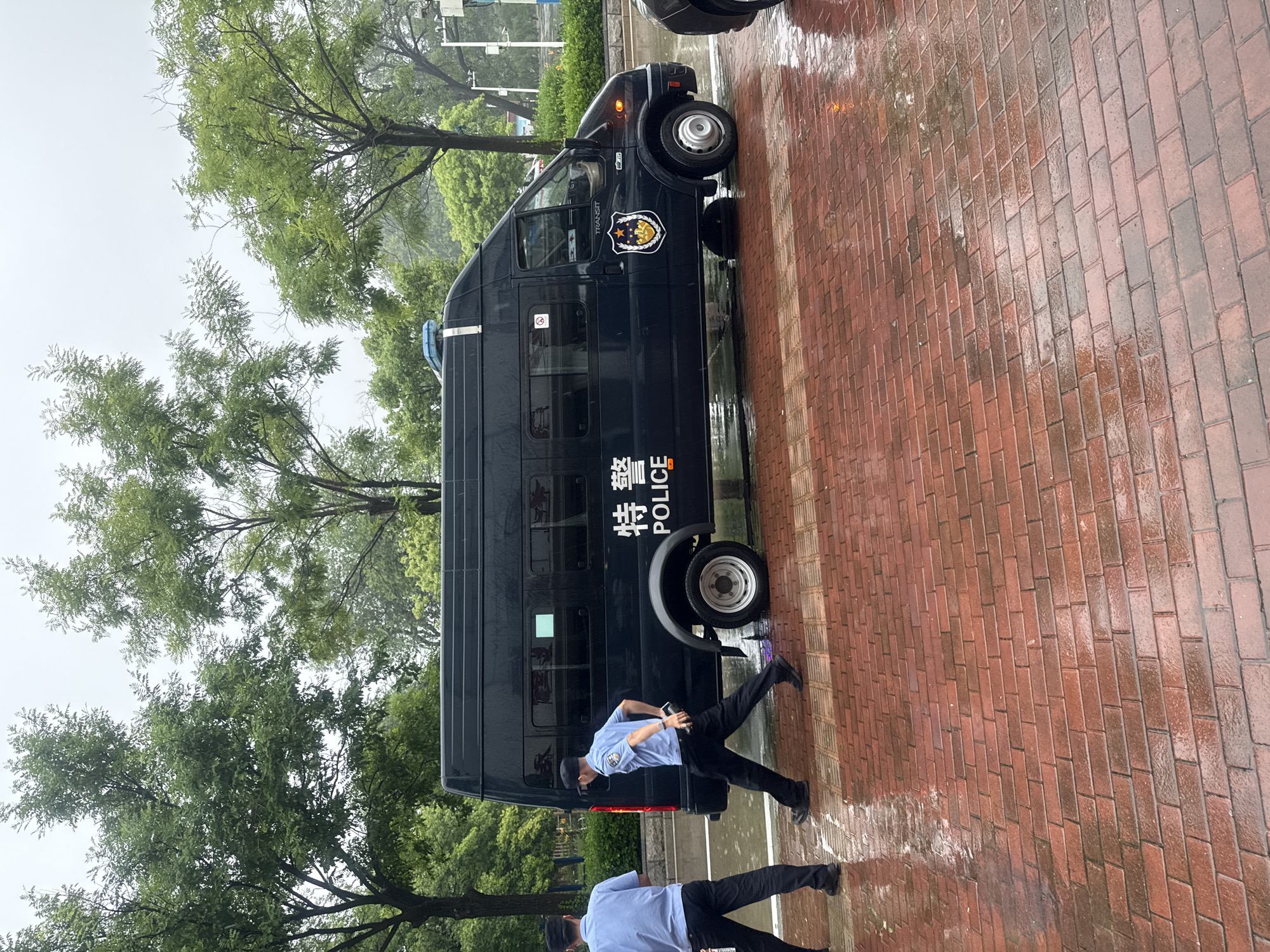 The image shows a black police van with Chinese characters and the word "POLICE" on its side. The van has a flashing blue light on its roof. Two police officers in light blue shirts and dark pants are walking near the van. The ground is paved with red bricks and is wet, indicating it is raining or has recently rained. Trees are visible in the background. The scene appears to be on a street in Qinhuangdao, China.