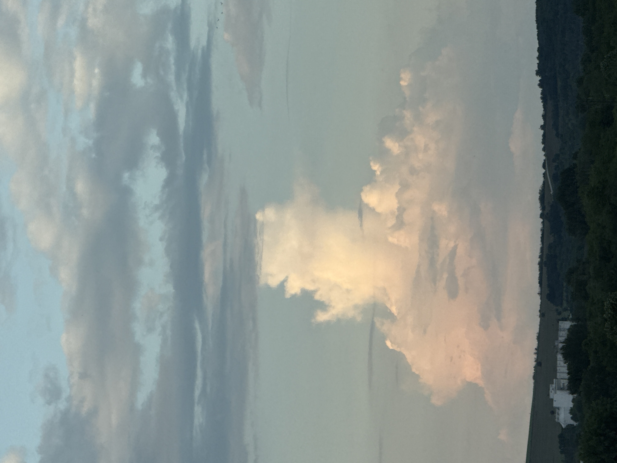 The image depicts a cloudy sky with a strip of wooded area and buildings at the far right. The sky is predominantly blue and gray with a large, bright, cloud formation tinged with yellow or pink light. The trees appear dark green. Several white buildings are visible amidst the trees at the bottom right corner. The image appears to be taken outdoors during daylight, with the landscape appearing in the lower right-hand corner. There is no information from the image to suggest its location in Lovech, Bulgaria.