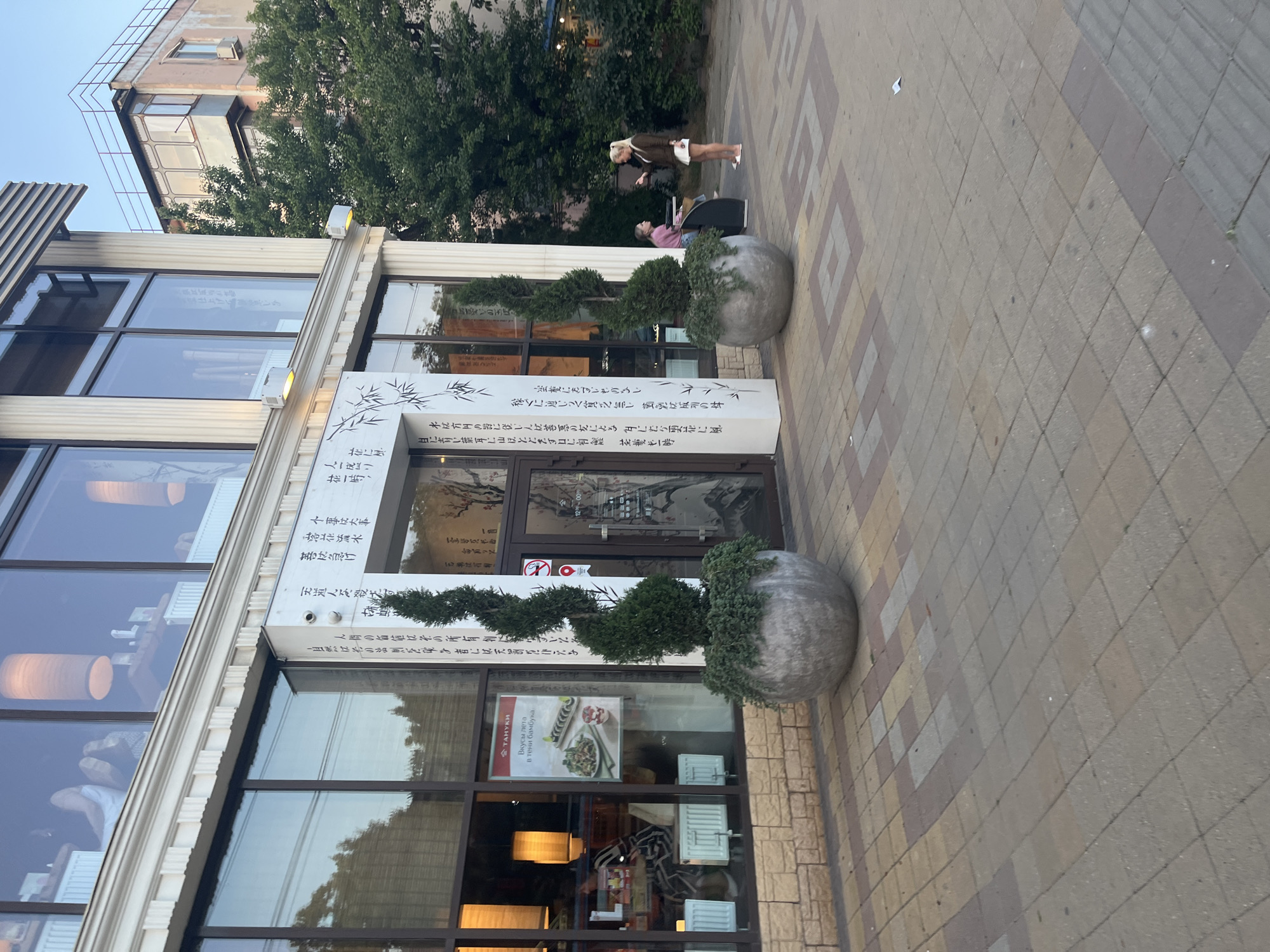 The image captures the exterior of a restaurant with Japanese-themed decor in Krasnodar, Russia. The restaurant features large windows, white decorative columns, and signage with Asian calligraphy. Two decorative spiral-shaped topiaries in large concrete planters flank the entrance. In the foreground, the pavement is made of square tiles in various shades of brown and beige, with a geometric pattern in places.
In the middle ground, a woman with blonde hair pulled back in a ponytail stands on the pavement, wearing a brown jacket and a white skirt. She is looking at her phone, which she holds in her right hand. To her left, a young woman with dark hair sits on a dark bench, also looking at her phone.
The background shows a building with a light pink facade and balconies, partially obscured by a large, mature green tree. The sky above is a clear, light blue. Inside the restaurant, visible through the windows, are patrons and decorative lighting. A poster advertising "Вкусы лета в тени бамбука" (Summer flavors in the shade of bamboo) is displayed on a window.