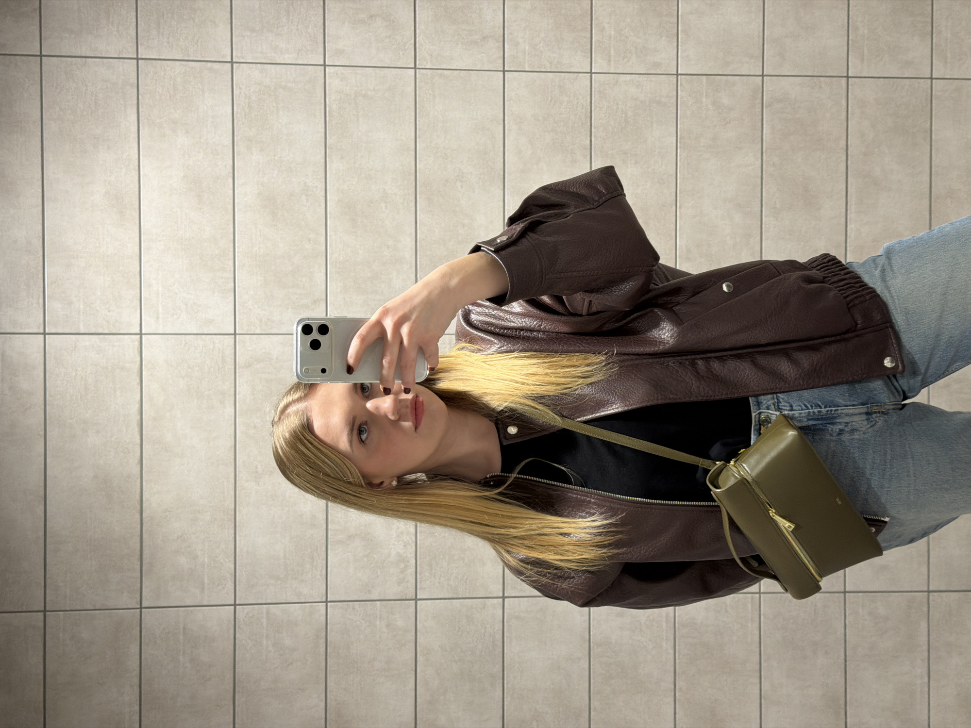 A woman with blonde hair is taking a selfie with a smartphone in front of a wall with beige square tiles. She's wearing a brown leather jacket, a black top, and light blue jeans. A small olive-green purse hangs on her shoulder. The exact location in Riga, Latvia, is not discernible from the visual information.