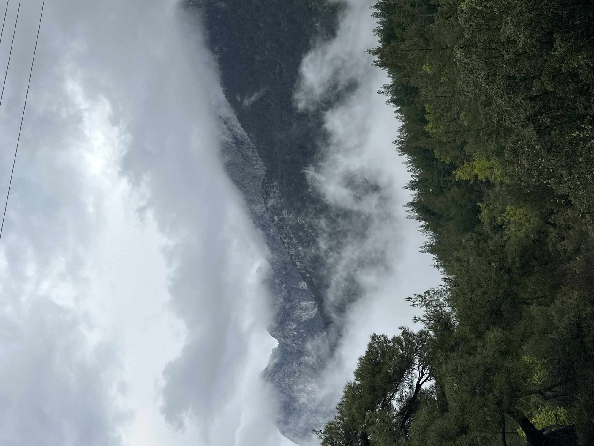 The image depicts a mountain shrouded in clouds, adjacent to a dense forest. The sky is overcast, indicating cloudy weather conditions. Power lines are visible in the top left corner of the image. The location is not discernible from the image alone, and there is no evidence it is in Konyaaltı, Turkey. No people, actions, or interactions are visible in the image.