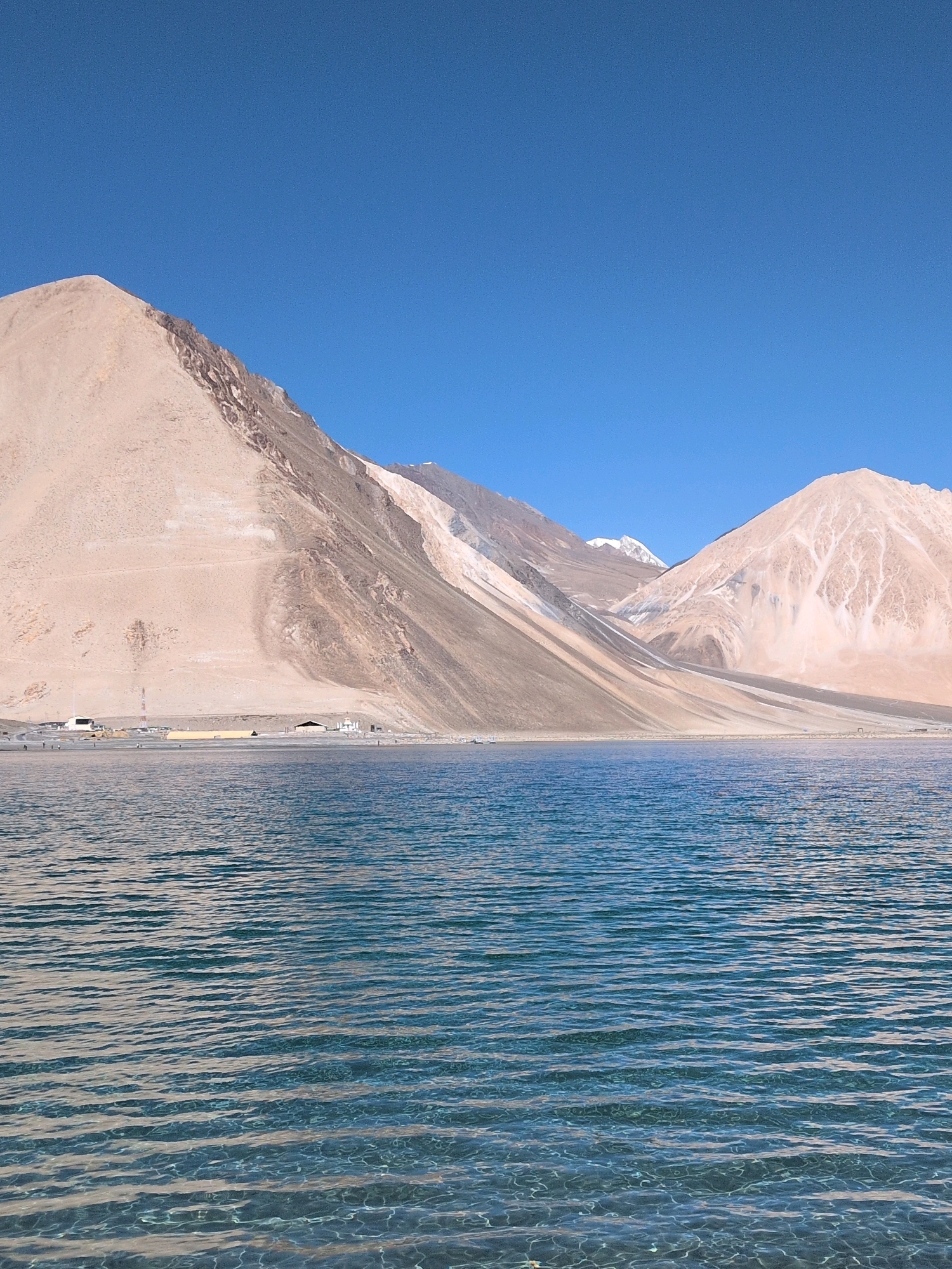 An expansive, clear blue sky stretches across the top of the frame, meeting a range of towering, barren mountains with sandy, ochre-colored slopes. The mountains are characterized by sharp, angular forms, with visible striations and patches of lighter and darker earth. Some distant peaks are capped with snow, contrasting with the dry, desert-like appearance of the lower slopes.
At the base of the mountains, a narrow, flat stretch of land is visible, dotted with several small, low-lying buildings and structures. Some appear to be simple dwellings or outposts, while others are more industrial, such as a tall communication tower. A few tiny figures of people can be seen scattered along this shoreline, suggesting a sparse human presence.
In the foreground, a large body of water occupies the lower half of the image. The water is a deep, vibrant blue, with a surface textured by numerous small, consistent ripples that catch the light. The clarity of the water allows for hints of the submerged surface to be seen beneath the ripples, creating a shimmering effect. The overall scene conveys a sense of vastness, natural beauty, and remote, arid landscape.