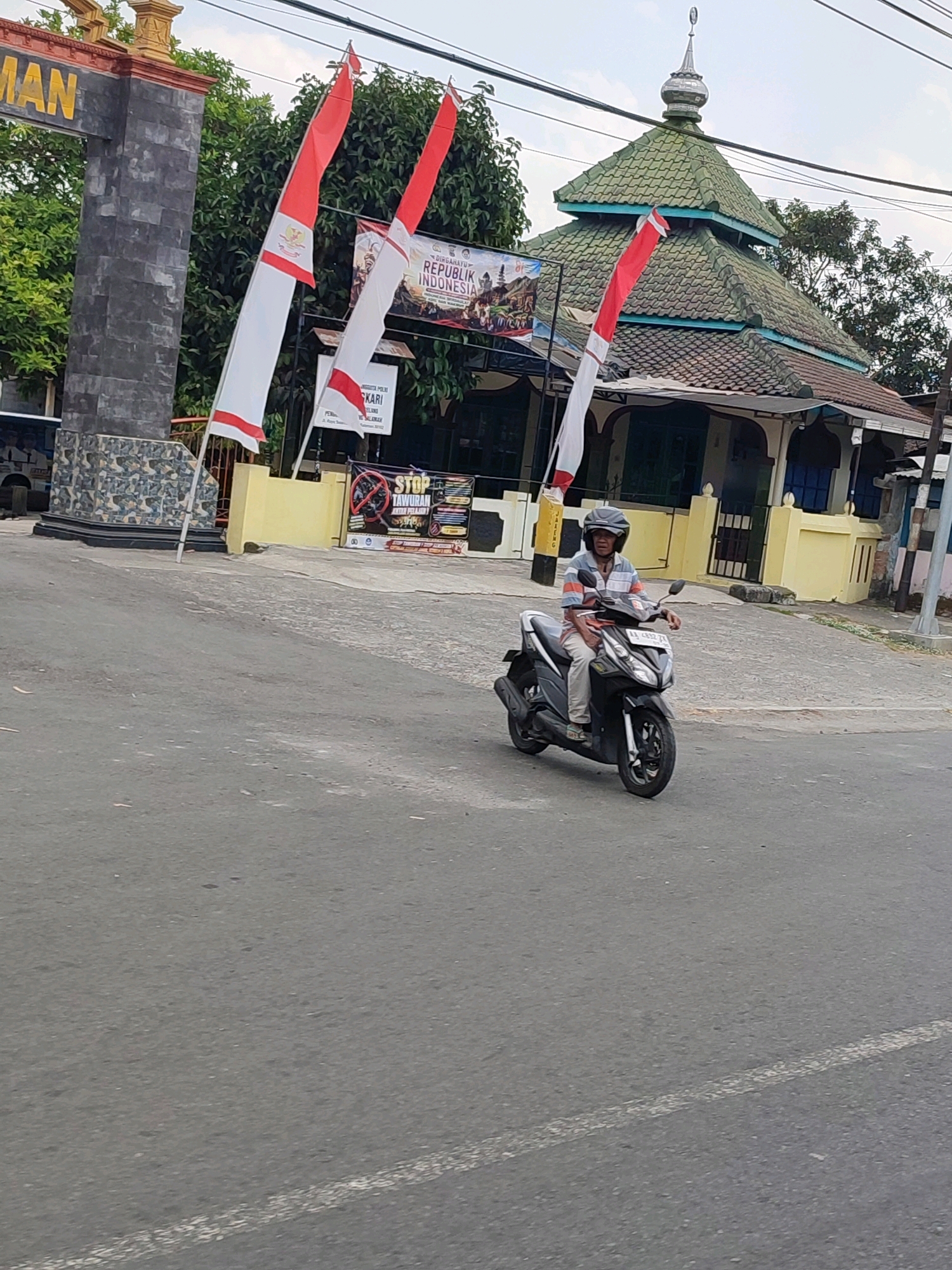 An elderly man wearing a helmet and a striped shirt rides a black scooter down a paved road. In the background, a mosque with a green tiled roof stands behind a yellow building. Several Indonesian flags, red and white, are planted in front of the mosque. A sign with text in Indonesian and an image of a group of people is also visible. The road has white lane markings. The scene appears to be in the city of Salaman, Indonesia.