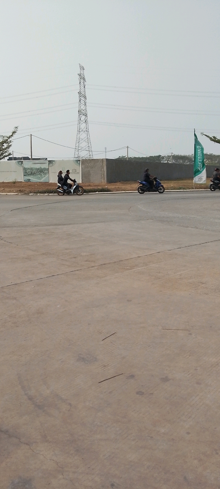 An outdoor, daytime scene in a developing area, possibly on the outskirts of Satriamekar, Indonesia. The foreground is dominated by a vast expanse of concrete pavement, with faint tire marks and cracks visible. In the middle ground, a road runs horizontally, with several motorbikes and riders. To the left, a motorbike carries two individuals, a male driver and a female passenger. Further to the right, another motorbike is ridden by a single individual. To the far right, the back of another motorbike and rider is partially visible. Behind the road, a low concrete wall separates the road from a construction site. Large banners, some depicting residential buildings, are mounted on the wall. A tall electricity pylon stands prominently in the background, with power lines stretching across the hazy sky. A green flag with white lettering and a logo is planted on the right side of the frame, near the edge of the construction site. The overall impression is one of ongoing development in a semi-urban or rural setting.