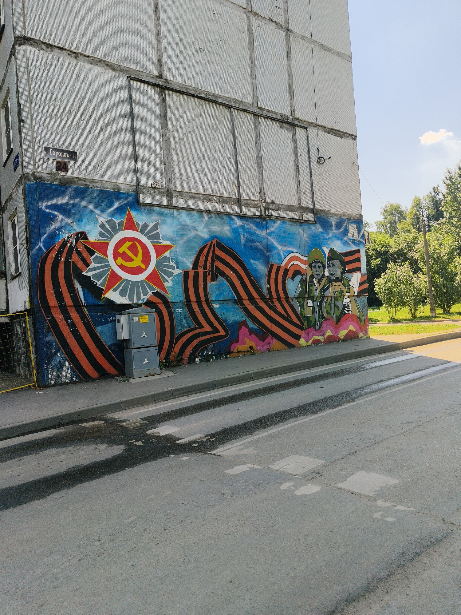 An outdoor, eye-level, medium shot captures a building with a mural painted on its exterior wall in an urban setting. The mural, which dominates the lower portion of the image, features a prominent red star with a yellow hammer and sickle emblem, symbolizing the Soviet Union. This emblem is flanked by what appear to be St. George's ribbons, characterized by their black and orange stripes, draped in a flowing manner. To the right of the emblem, two figures in military attire are depicted. The building itself is a multi-story structure with a facade covered in small, square, light-colored tiles, and vertical dark lines dividing sections of the wall. Above the mural, a street sign reading "ул. Городок" (Gorodok Street) is visible, along with a building number "24". In front of the mural, there are two gray electrical boxes stacked atop one another. The street in the foreground is asphalt, with painted white crosswalk lines and visible water stains. The background shows a grassy area with trees and a clear blue sky with a single white cloud. The overall scene suggests a public space, possibly a residential area, adorned with historical or patriotic artwork.