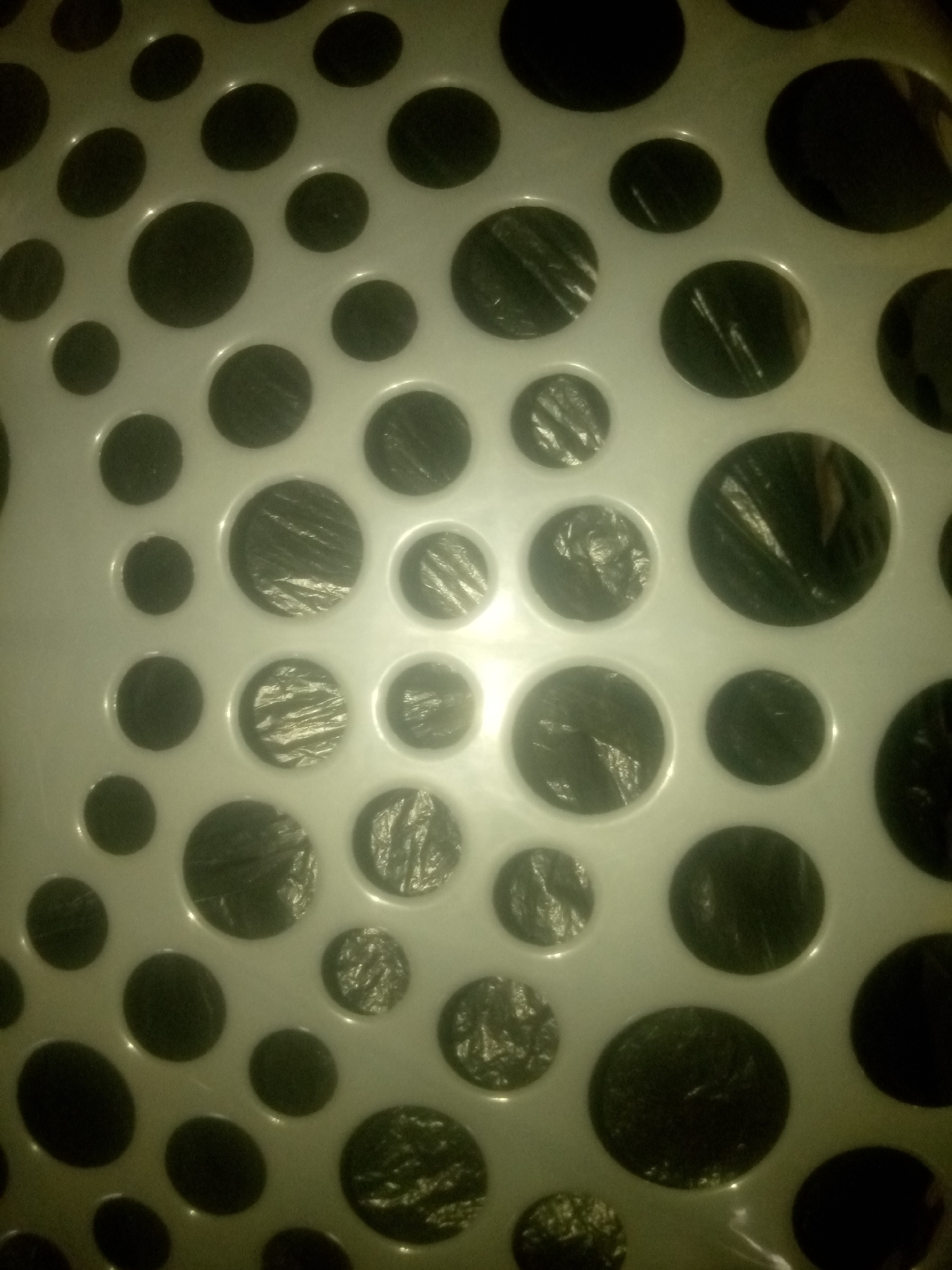 The image is a close-up, abstract shot of a white surface with numerous circular indentations, many of which are filled with dark material that appears to be plastic sheeting. The lighting creates a dappled effect, with some areas brightly lit and others in shadow. The texture of the plastic sheeting within the indentations is visible, showing a crinkled or wrinkled pattern. The overall impression is of a perforated material or a container with a patterned bottom. No specific location in Jauro Yinu, Nigeria can be determined from this image.