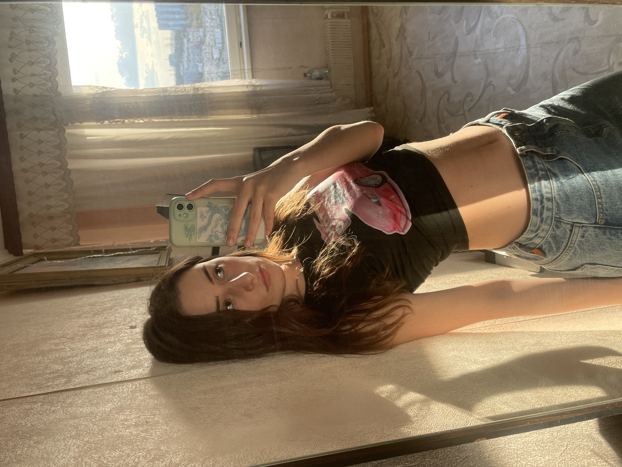 A young woman with long dark brown hair is lying on a light-colored, possibly reflective surface, possibly a table or a mirror, and taking a selfie with a green smartphone. She is wearing a short black t-shirt with a pink and red graphic design on the front, and high-waisted light blue jeans. Her midriff is visible. The background shows a window with light-colored curtains. Beyond the window is a cityscape, suggesting a view from an apartment building in an urban area. Based on the architectural details of the buildings, it is possible this is a location in the city of Moscow, Russia. In the upper right corner of the frame is a radiator.
