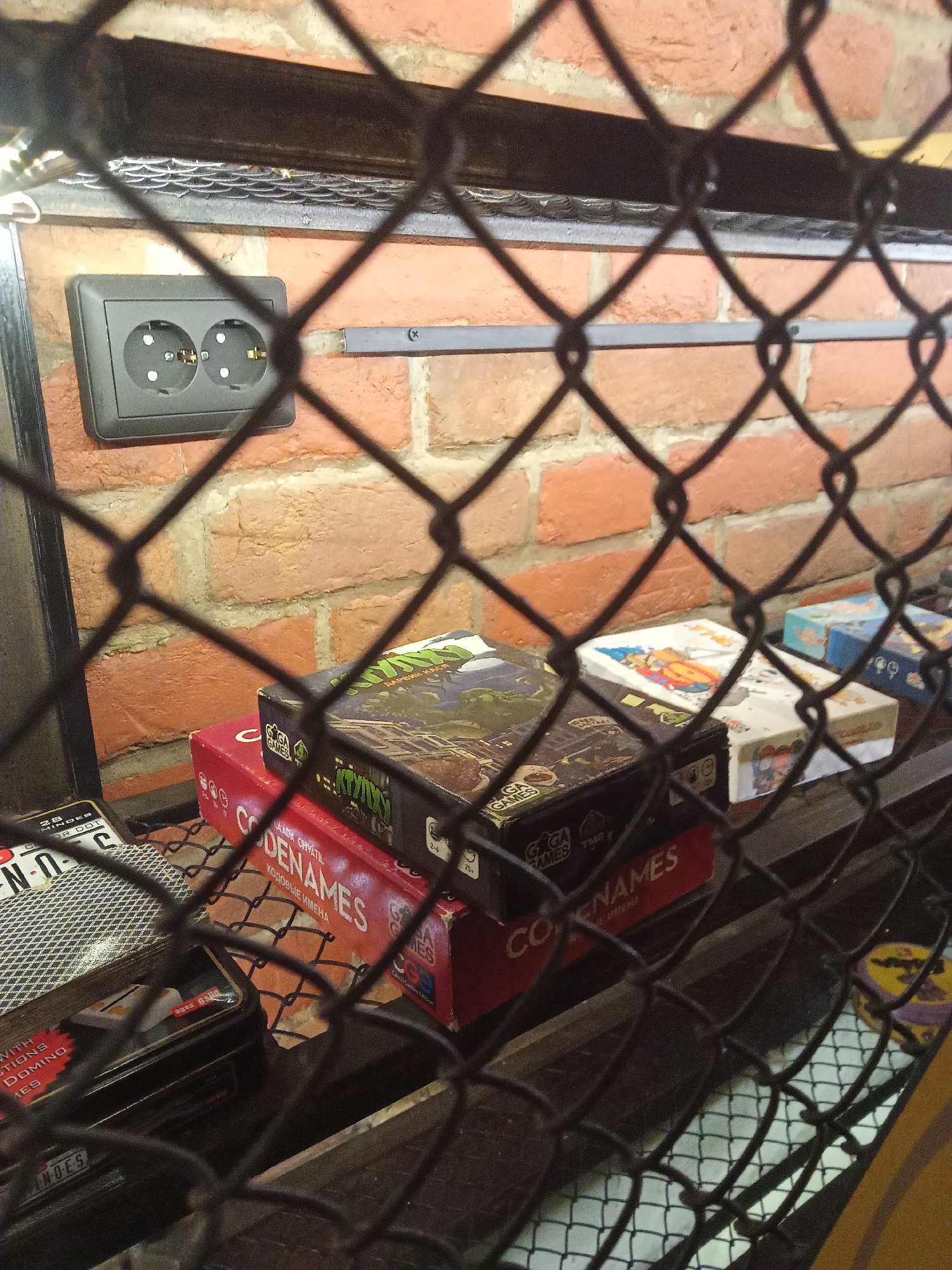 A close-up, slightly angled shot looks through a black metal chain-link fence at a brick wall, a black electrical outlet, and several board games. The electrical outlet has two circular openings and a grounding pin slot in each. It is mounted on a red brick wall. Below the outlet, on a shelf, are several board game boxes. Two prominent boxes are red with white lettering that reads "CODENAMES" and a green box with colorful illustrations. Other board game boxes, some partially obscured, are visible to the right. The lighting appears to be artificial and warm, casting some shadows. The scene is indoors, and the chain-link fence creates a layered, partially obscured view.