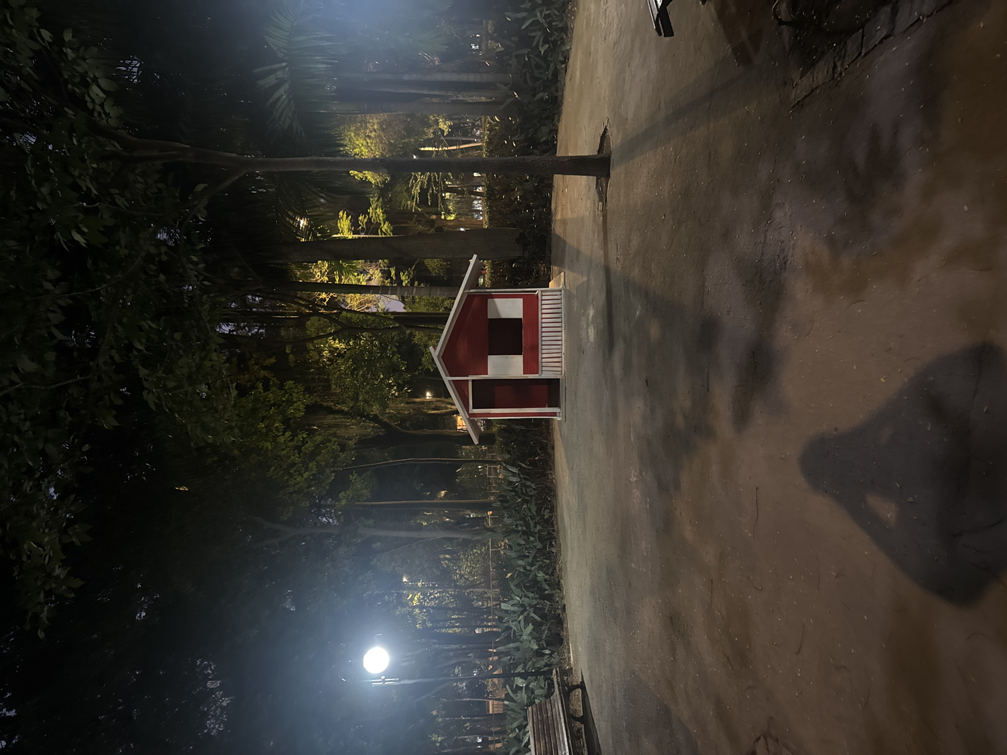 This is a night-time photograph taken in the Parque da Cidade in Niterói, Brazil. The scene is dimly lit by a street lamp on the left, casting a bright glow on the path and surrounding trees. In the center of the image, a small, red and white shed with a peaked roof is visible. To the left of the shed, a wooden park bench is partially visible. The ground is a mixture of dirt and asphalt, with some scattered leaves and debris. A prominent shadow of a person holding what appears to be a camera is cast on the ground in the foreground. The background is filled with dense foliage and tall trees, some of which are illuminated by distant lights. The overall atmosphere is serene and quiet, typical of a park at night.