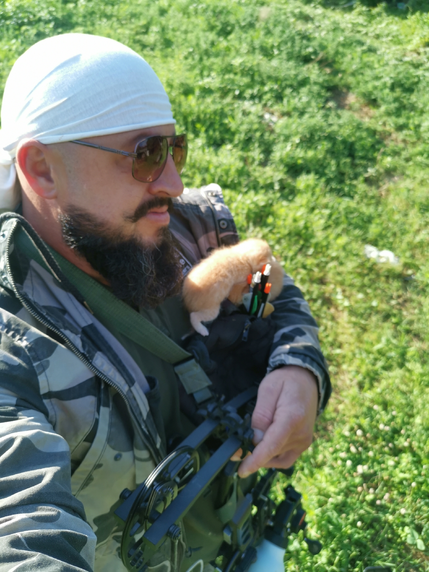 A man wearing a white bandana, sunglasses, and a camouflage jacket is holding a kitten while standing in a grassy area. The kitten, which appears to be ginger and white, is nestled in a black pouch attached to a bow. The man has a beard and mustache. The environment is outdoors with green grass and a bright sky. The location is identified as Semypolky, Ukraine.