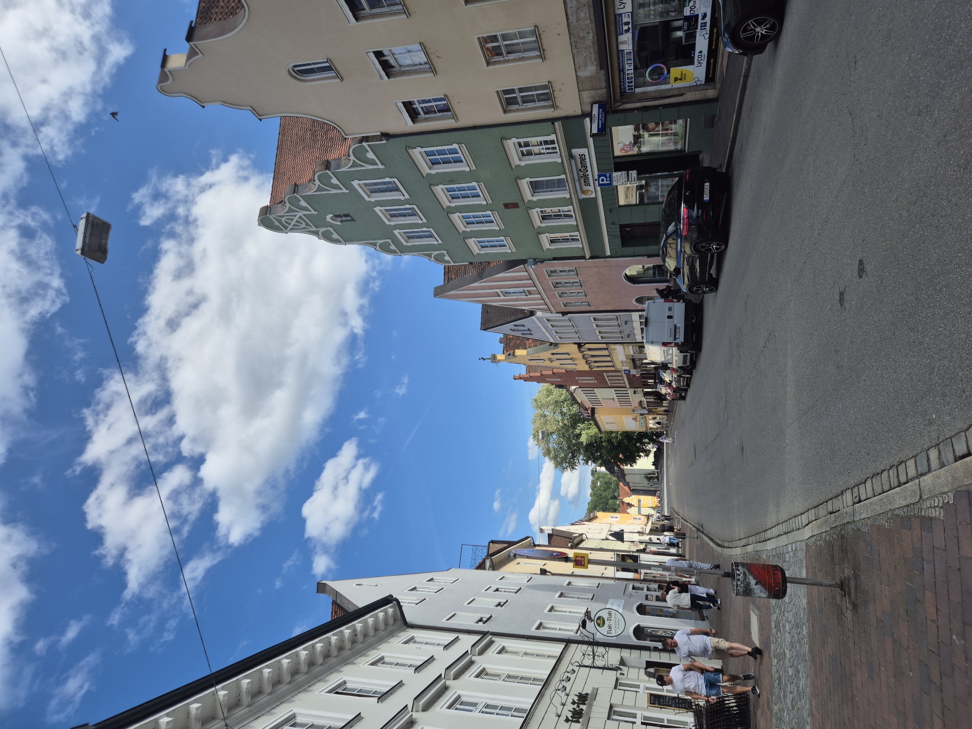 The image depicts a street scene in Landshut, Germany, under a bright blue sky with scattered white clouds. The street is lined with colorful buildings exhibiting a traditional European architectural style, characterized by their pitched roofs and ornate facades. On the left side of the street, a row of lighter-colored buildings with multiple windows is visible, some with awnings and signage for businesses. Further down, the buildings become more varied in color, including a prominent green building with "smileGames" signage and a parking symbol. On the right side, a series of buildings in shades of beige and yellow are visible, also with shops on the ground floor. The street itself is paved with asphalt and bordered by sidewalks. Several cars are parked along the right side of the street, and a few people are walking on the sidewalks. One person is seen pushing a stroller, and another is walking a dog. In the foreground on the left sidewalk, a trash can with red and white markings is positioned near two men walking. The scene appears to be a typical European town street on a sunny day.