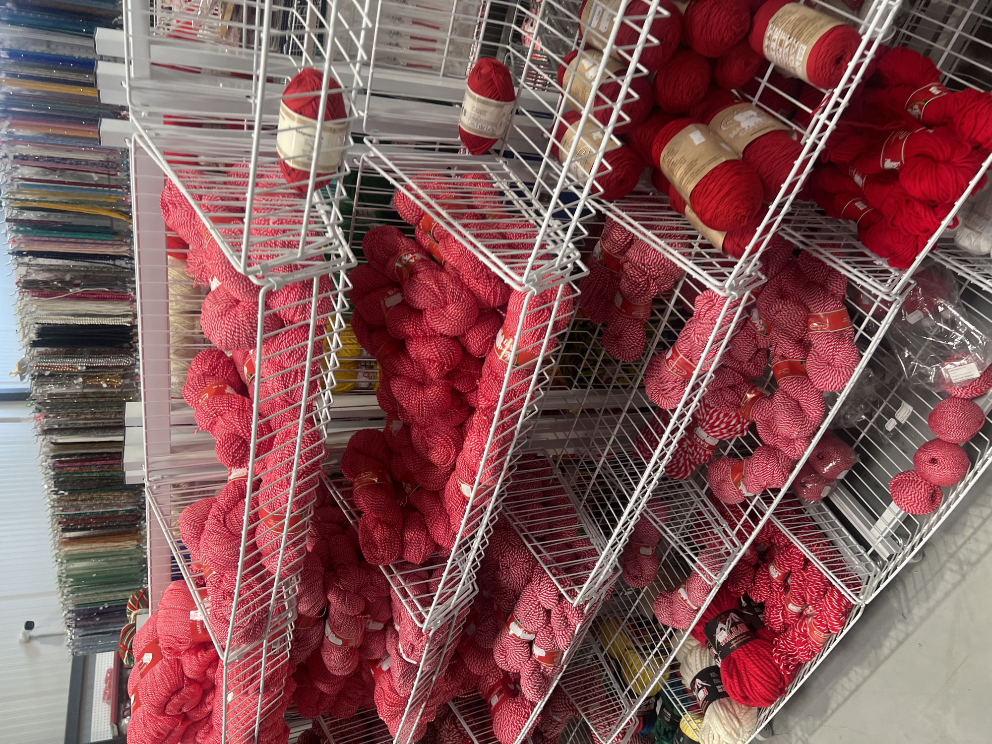 The image shows a retail display of yarn spools in what appears to be a craft store. The yarn spools are various shades of red, and some have white accents. They are arranged in multiple white wire shelves. To the left of the shelves, there is a wall display of fabric or ribbons. The location is a store in Burgas, Bulgaria.
