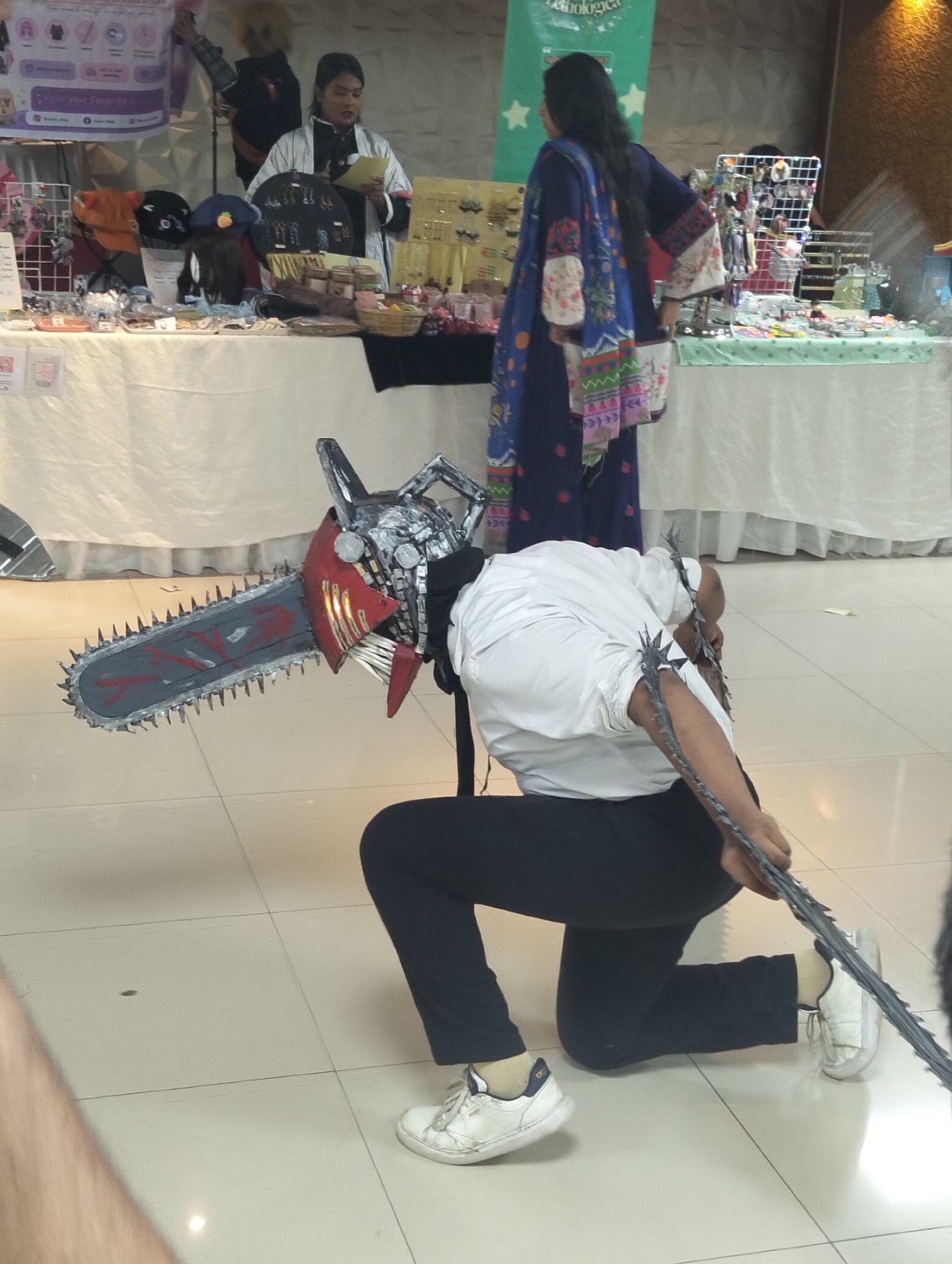 In an indoor, tiled environment, a person dressed in a white shirt and black pants is kneeling on the floor. They are wearing white sneakers and have a costume on that includes a large chainsaw prop. The prop is red and gray with a spiked chain. The person's face is obscured by the chainsaw mask, and their arms are raised, holding the prop. In the background, several individuals are standing behind tables laden with merchandise. One woman is wearing a light-colored robe and holding a paper, while another person in a dark blue outfit with a colorful patterned shawl is standing to the right. The tables display various items, including hats, jewelry, and other products. The overall scene appears to be a convention or market event.