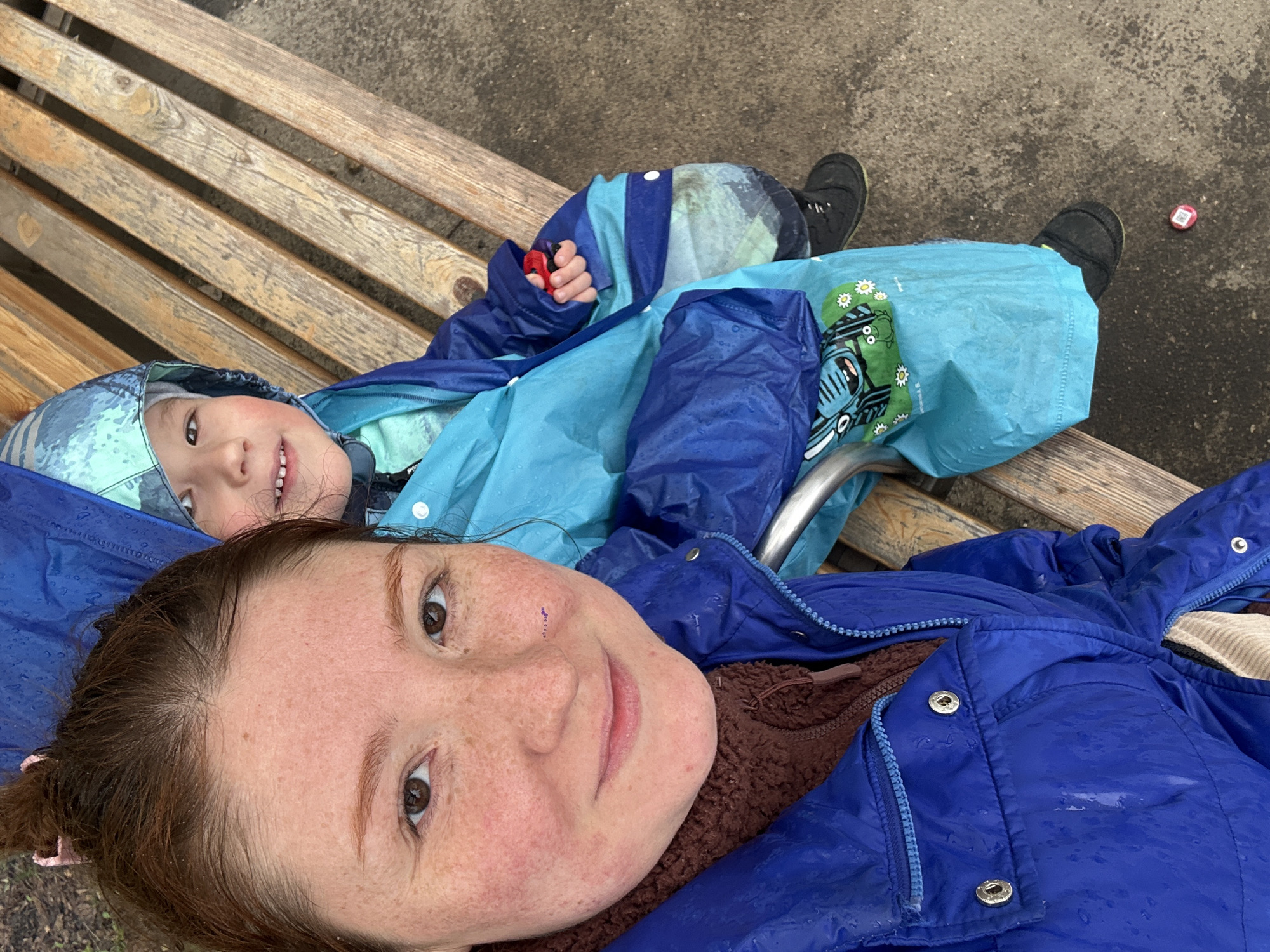 A woman and a child are lying on a wooden bench outdoors. The woman, facing the camera, has reddish-brown hair pulled back and is wearing a blue jacket over a brown fleece. Her face is freckled. The child, positioned above her, is wearing a light blue and dark blue hooded waterproof suit and black shoes. The suit has cartoon train imagery on it. The child is holding a small red object in their hand. The bench is made of light-colored wooden slats. The ground next to the bench is a mix of concrete and dirt. There is a red and white object next to the child's shoes on the ground. Water droplets appear to be on their jackets. The location is Nizhny Novgorod, Russia.