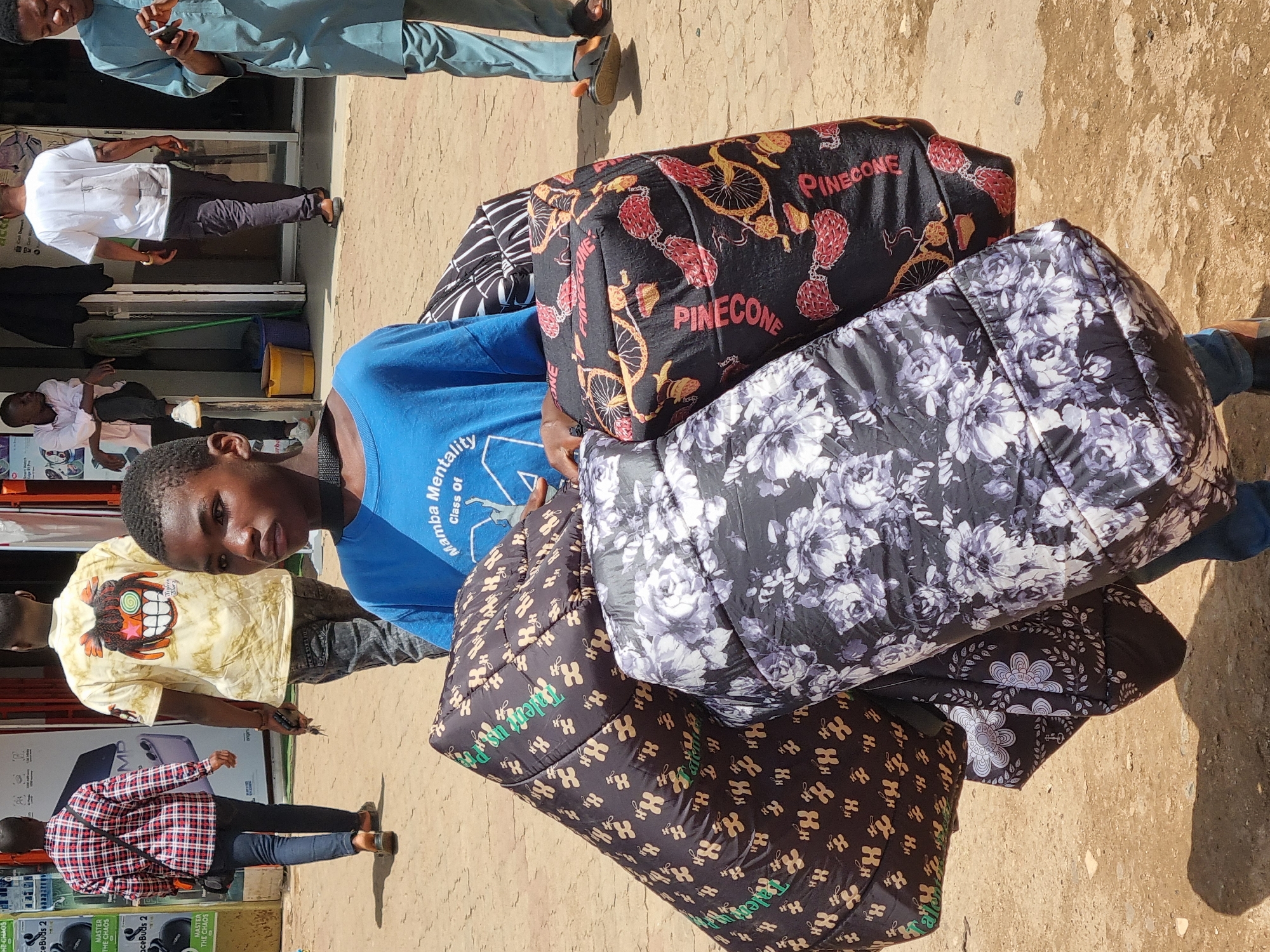 A young boy, appearing to be in his early teens, is the central subject of the image, carrying a substantial load of what appear to be rolled-up sleeping bags or thick blankets. He is positioned in the foreground, facing slightly to the right. He wears a blue t-shirt with white text that includes "Mamba Mentality" and "Class Of". His expression is neutral.
Behind him, several other individuals are visible in the background, suggesting an outdoor market or street scene in a city. To the left, a person wearing a red and black checkered shirt and blue jeans walks away from the camera, carrying a dark-colored bag over their shoulder. Further back, another individual in a light-colored t-shirt with a graphic design on the back is walking. To the right, a person dressed in a light blue traditional outfit is walking with a phone in hand.
The environment appears to be an unpaved street or pathway, with sand and dirt covering the ground. In the background, buildings with storefronts are visible, suggesting a commercial area. The lighting indicates it is daytime, with sunlight casting shadows on the ground. This scene is consistent with the environment of Jos, Nigeria, known for its bustling markets and vibrant street life. The overall impression is of a busy urban setting where commerce and daily life intersect.