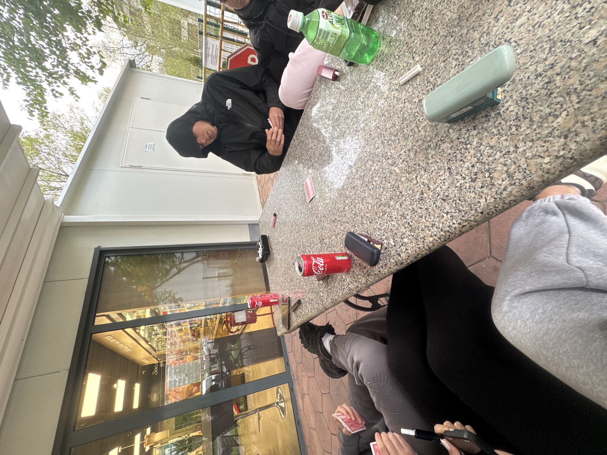 The image captures an outdoor gathering around a speckled granite-like table during the daytime, viewed from a low and tilted angle. Three individuals are visible. One person, wearing a black hooded "The North Face" jacket, sits on the left side of the table, focused on playing cards. Another person is partially seen behind them in a dark jacket. On the right, a third individual is visible from the waist down, wearing grey sweatpants and black leggings, holding playing cards and a smartphone with white fingernails.
The table is adorned with several items, including two red "Coca-Cola" cans, a green bottle of "Aloe" drink, a light green eyeglasses case, a lighter, a white cylindrical object, and scattered red-backed playing cards.
In the background, to the left, stands a white modular building, possibly a kiosk or portable cabin, with a door. Above this structure, green trees and a bright sky are visible. To the right, a large glass window reflects the interior of what appears to be a shop or cafe, showing shelves, lights, and furniture. Visible text in the reflection includes "3aC" and "cafe" in Cyrillic, suggesting the location uses the Cyrillic alphabet. The ground is paved with reddish tiles.
The activity appears to be a casual card game among friends, enjoying beverages in an informal setting. The overall mood seems relaxed.