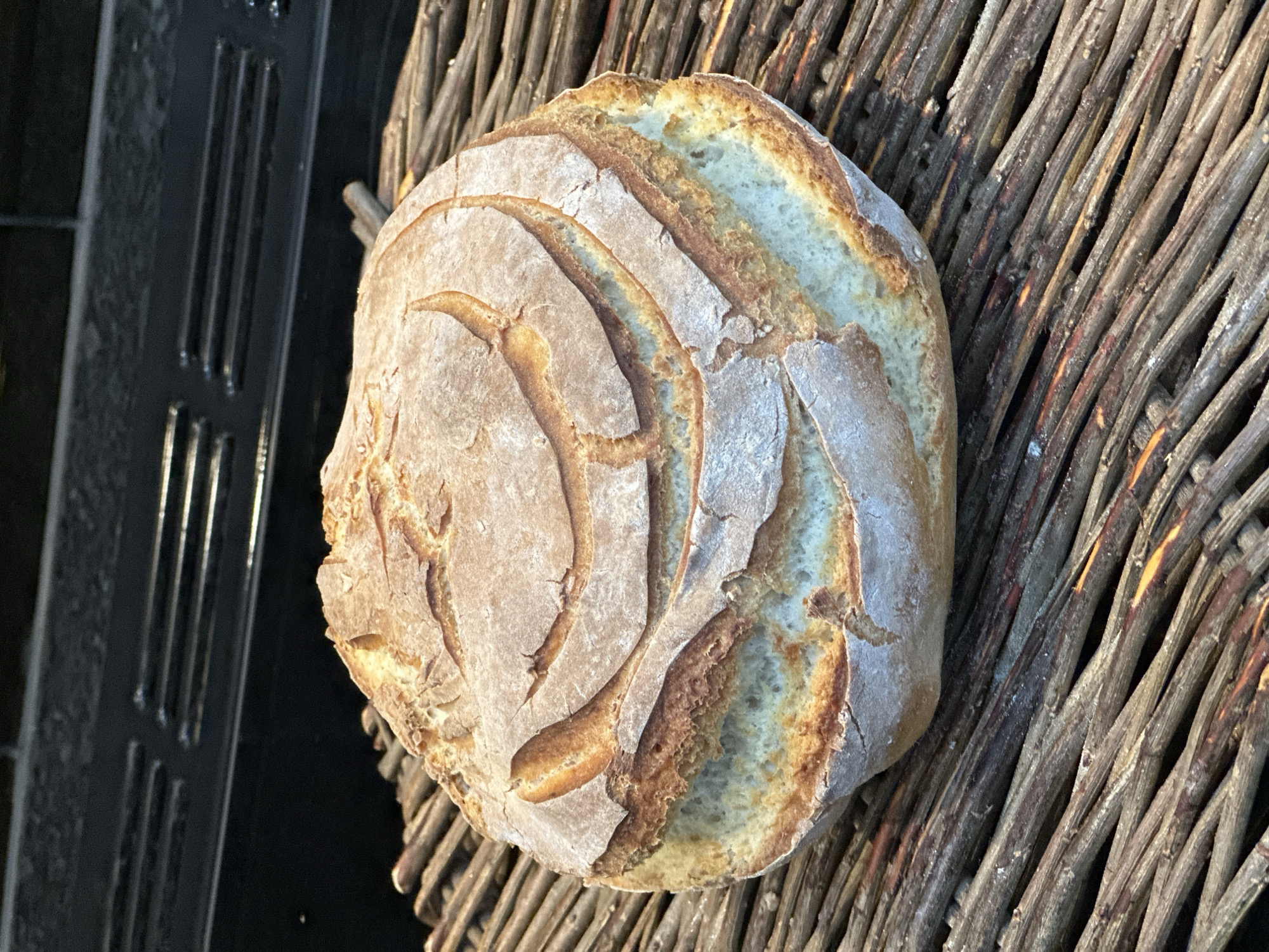 A loaf of artisanal bread rests on a woven twig surface, possibly a basket or placemat. The bread is round with a textured crust featuring decorative scoring marks. The crust is golden brown, transitioning to a paler, almost white hue around the edges, suggesting a light dusting of flour. The backdrop is a combination of the woven twigs and a black, possibly metallic, surface with vertical slots on the left, hinting at a shop display or shelf. The lighting is natural, casting shadows and highlighting the textures of both the bread and the twigs. This scene could be from a bakery or market in Tunbridge Wells, United Kingdom, based on the style of bread and the rustic presentation. There are no people visible.