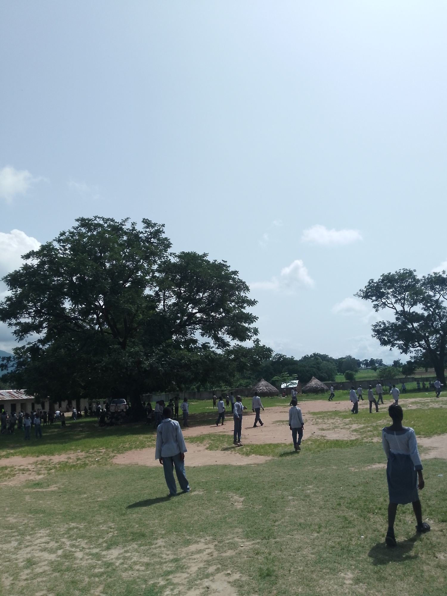 The image depicts an outdoor scene in Serti, Nigeria, likely a schoolyard, with a large, leafy tree dominating the left side of the frame. A group of people, appearing to be students in uniform, are scattered across a grassy field, some walking and others potentially engaged in activities. In the background, there are several thatched-roof structures and buildings, suggesting a rural or semi-rural setting. A light-colored vehicle is parked near one of the buildings. The sky is bright and clear with a few scattered clouds. The overall impression is of a daytime gathering or event taking place in a community space.