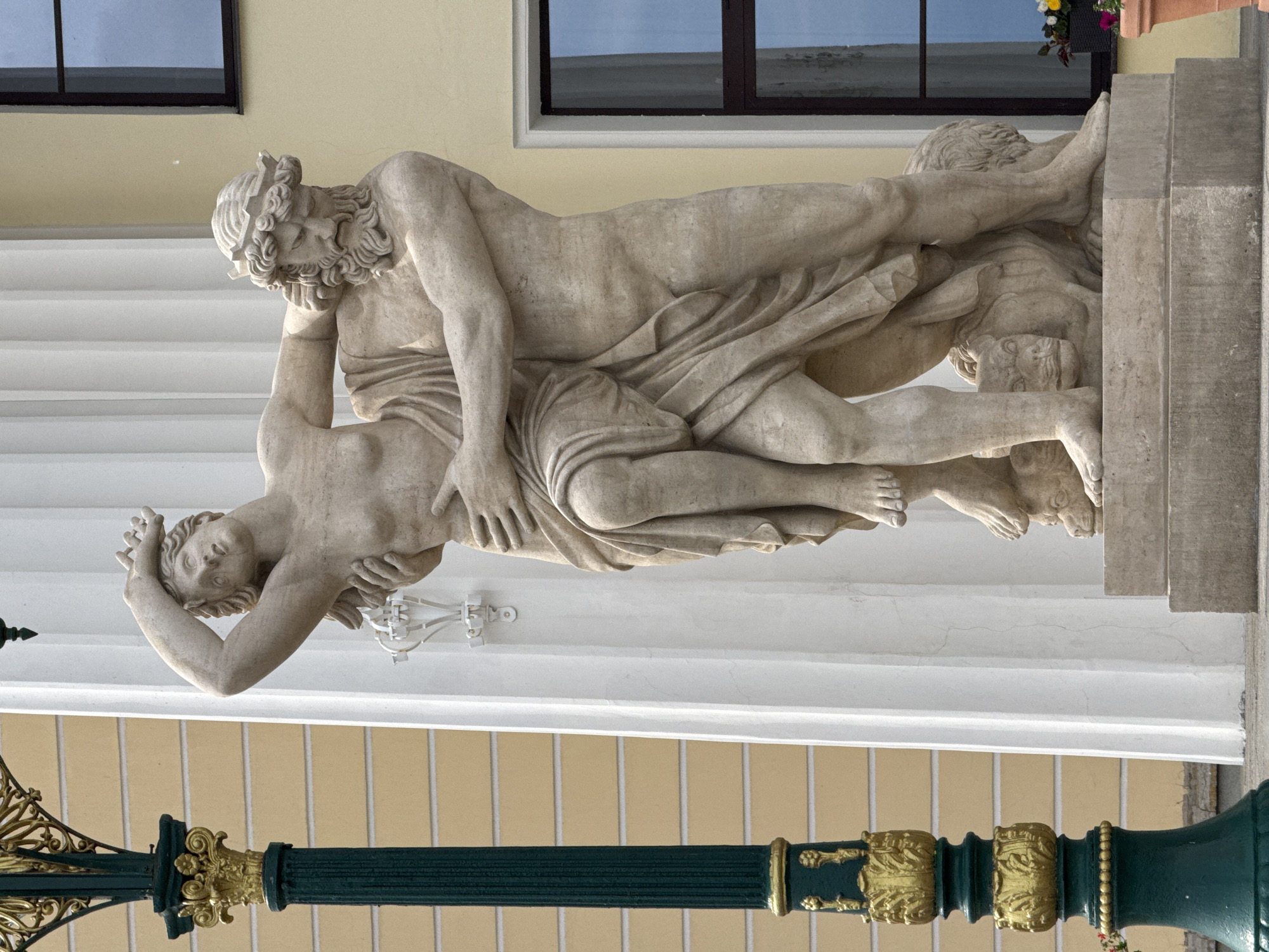 The image shows a close-up of a stone sculpture depicting a man and a woman, with the man embracing the woman. The sculpture is situated outdoors, with a light yellow building and a tall green lamppost with gold accents visible in the background. The woman is depicted with her arm raised and her body partially covered by drapery, while the man has a beard and is wearing a crown of leaves. Both figures appear to be standing on a stone pedestal that supports a lion's body. This sculpture is likely a detail from the exterior of the Russian State Museum in Saint Petersburg, Russia, given its architectural style and placement against a neoclassical building.
