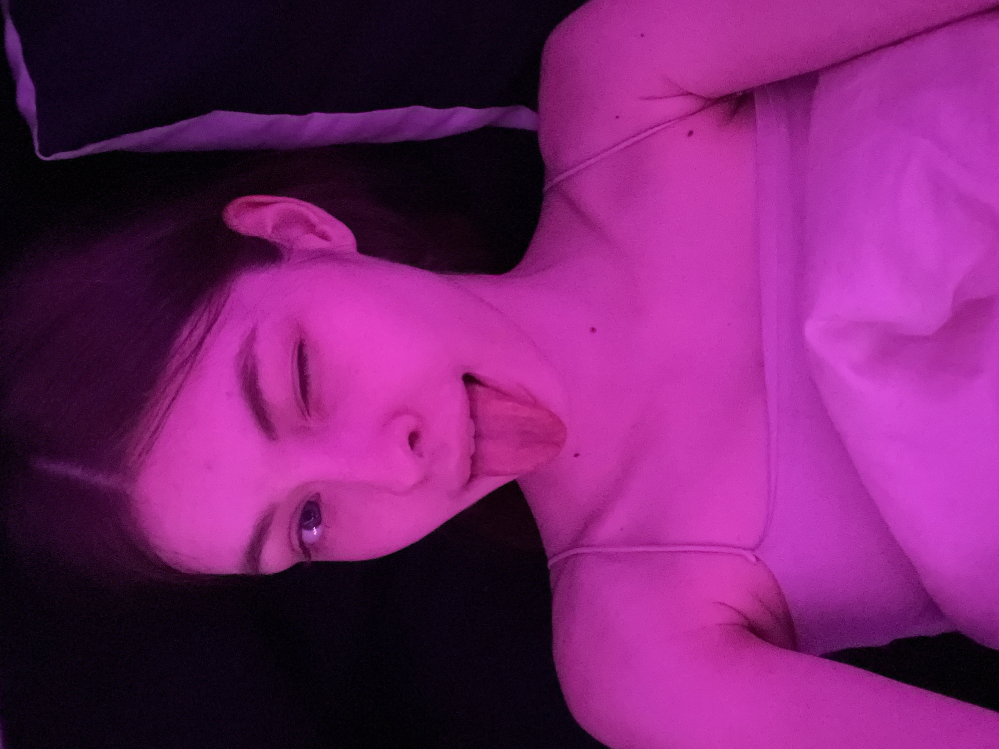 A young woman lies on a dark surface, possibly a bed, with a white pillow and a dark pillow nearby. She is winking and sticking out her tongue, and appears to be wearing a light-colored tank top or camisole. The entire scene is lit with a strong pink/purple light, which affects the colors of the skin, bedding, and surrounding area. The image does not contain sufficient information to determine if it was taken in Saint Petersburg, Russia.