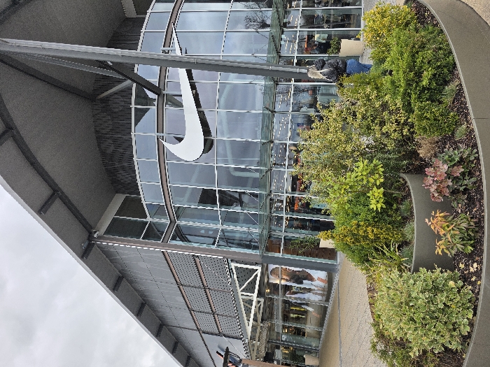 This image captures the exterior of a Nike retail store located within an outdoor shopping complex in Zweibrücken, Germany, specifically identifying with the Zweibrücken Fashion Outlet. The environment is depicted under an overcast sky.
In the foreground, a large, curved, multi-tiered concrete planter is filled with a variety of shrubs and small plants, exhibiting green, yellow-green, and reddish-brown foliage. The ground beneath is paved with light-colored pavers.
The dominant structure is a modern commercial building featuring a curved glass facade on its lower and middle sections. A large, white Nike swoosh logo is prominently displayed centrally on the glass. The upper portion of the building showcases a dark, patterned wall texture and is topped by a wide, grey, angled roof supported by exposed dark grey steel beams and structural elements. Through the glass, interior lighting, merchandise displays, and a large graphic image of a person are discernible.
One individual is visible on the right side of the image, standing near the building's facade and facing towards its entrance. This person has blonde, shoulder-length hair and is wearing a black puffer jacket and blue jeans. Additional blurred figures are observable inside the store through the glass windows. Two light-colored, cylindrical potted plants are positioned near the building's facade on the right.