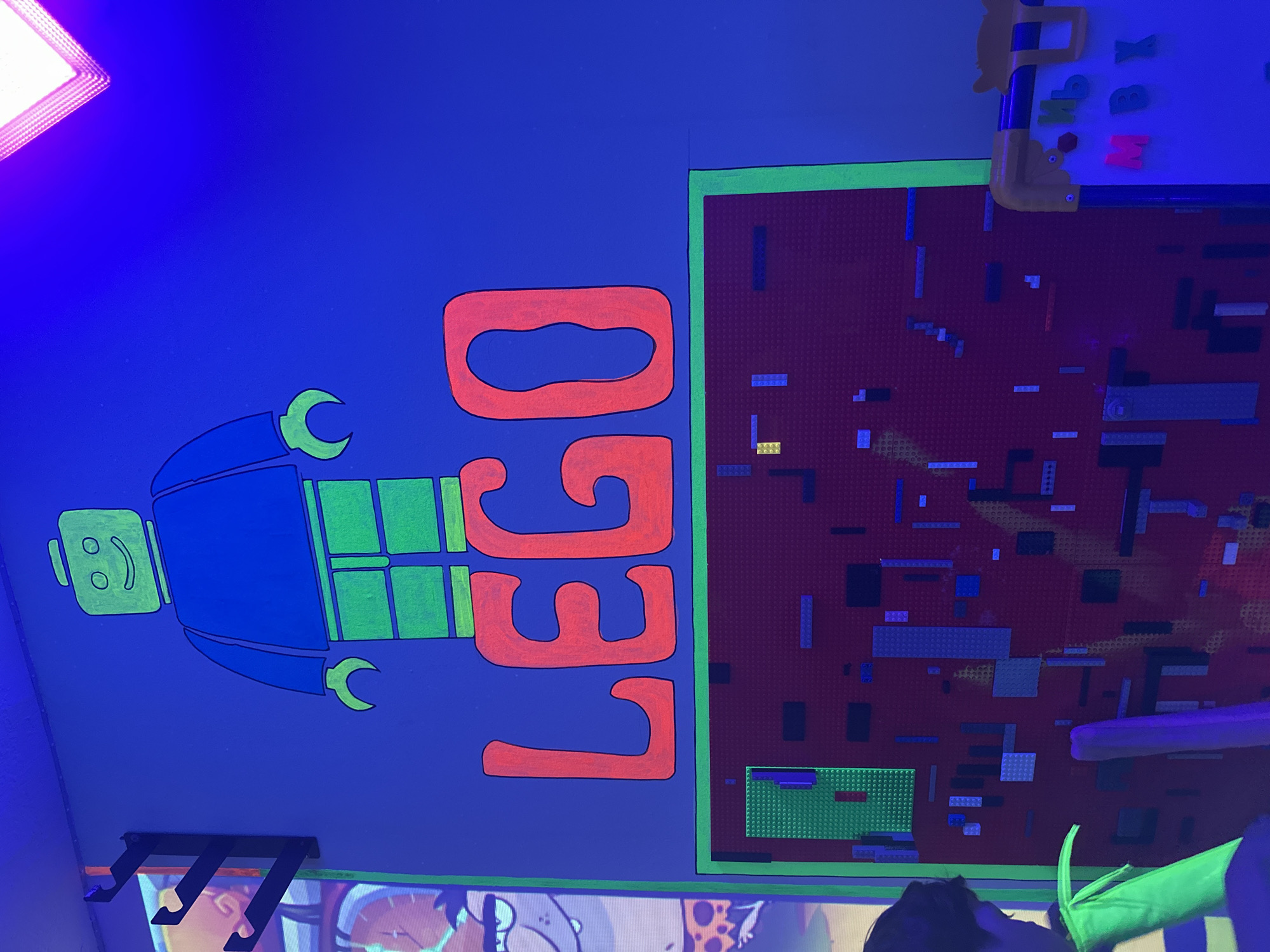 The image depicts a room featuring a blue wall with a painted Lego figure and the word "LEGO" in red with green outlines. A red Lego baseplate with various Lego bricks attached to it is affixed to the wall. The environment appears to be illuminated by a blue light, possibly UV. In the top right corner, part of a children's magnetic drawing board with colorful letters attached is visible. At the bottom of the frame, parts of cartoon characters are visible on another surface or wall. Three black coat hooks are positioned below the Lego figure. The location is Unknown, Unknown.
