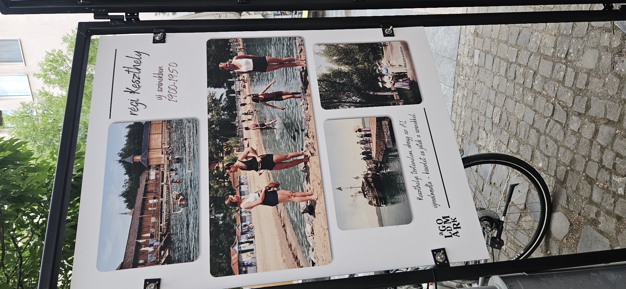 The image displays an outdoor exhibition showcasing historical photographs of Keszthely, Hungary, dating from 1900-1950. The exhibition is mounted on a white board supported by a black metal frame. In the foreground, the rear wheel and part of the frame of a bicycle are visible, leaning against the exhibition board. The ground is paved with cobblestones.
The exhibition board is divided into several sections. The top section features a panoramic view of a bathing establishment with wooden structures and people swimming in the water, captioned "1900-as évek eleje Szigetfürdő" (Early 1900s Szigetfürdő). To the right of this photograph, in large script, is the title "régi Keszthely" (Old Keszthely), followed by "új színekben" (in new colors) and the date range "1900-1950".
Below the top section, there are three smaller photographs. The left photograph depicts a beach scene with people in vintage swimwear wading in the water and sunbathing. The middle photograph is a close-up of a couple in bathing suits standing on the shore, with other people and beach huts in the background. This photograph is captioned "1935 Strandélet" (1935 Beach life). The right photograph shows a group of women dressed in long skirts and hats walking along a tree-lined path, captioned "1916 Séta parkban" (1916 Walk in the park).
A central photograph below the beach scenes shows a steamship docked at a pier, with people gathered on the pier and on the ship. This photograph is captioned "1911 Indul a hajó" (1911 The ship departs).
At the bottom left corner of the exhibition board is a logo that reads "AGO LDM ARK". Below the photographs, there is text that translates to "Keszthely's history, the AI has recreated it - a visit and play with colors." The background suggests an urban setting with greenery from trees and parts of buildings visible.