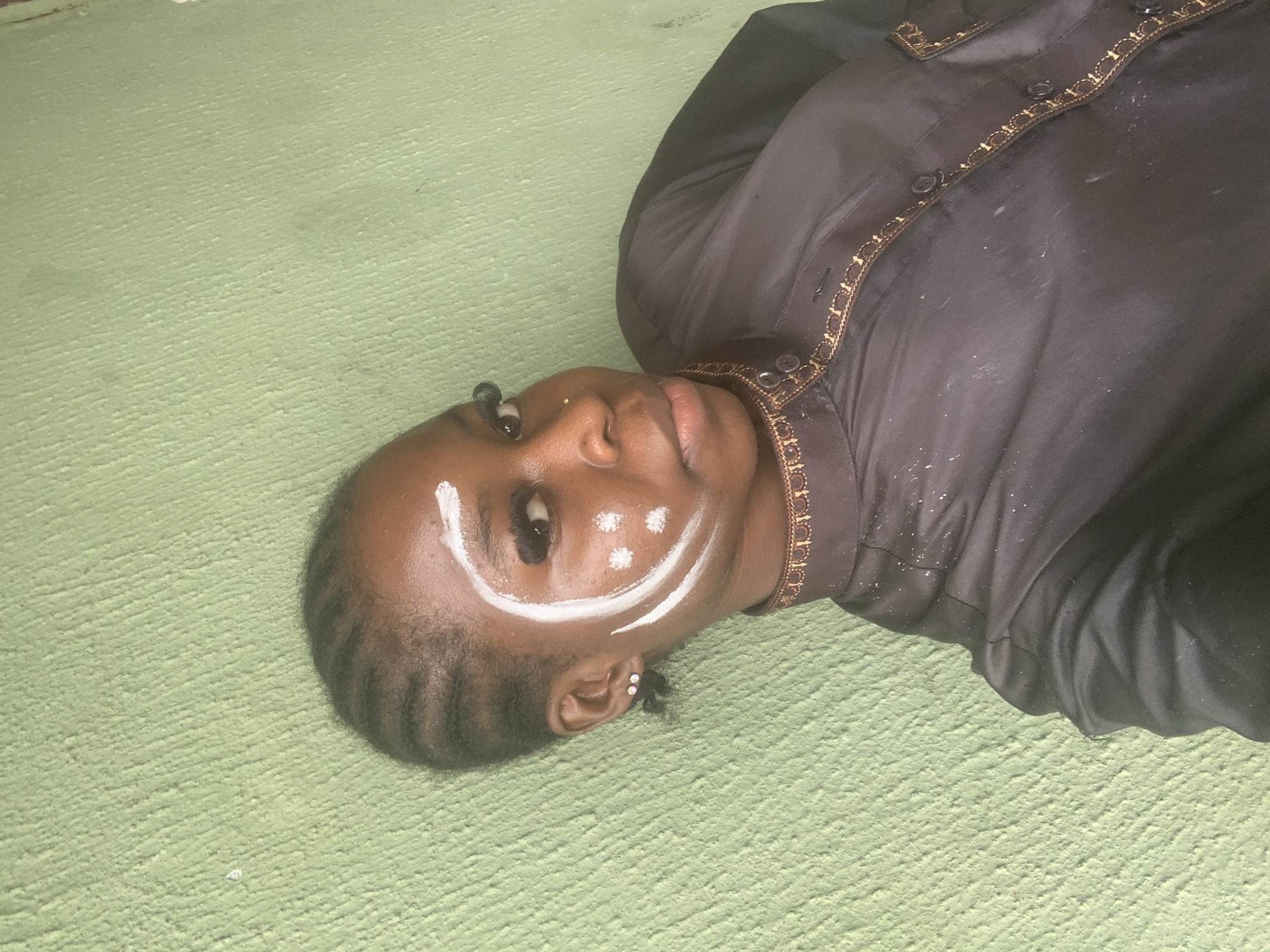 In Ado, Nigeria, a young Black individual with braided hair is positioned against a light green, textured wall. They are looking at the camera, with white face paint applied in a crescent moon shape around their right eye, accompanied by three white dots on their cheek. The individual is wearing a black garment adorned with gold trim around the collar and down the front, where buttons are visible. The wall exhibits some dust or debris scattered across its surface.