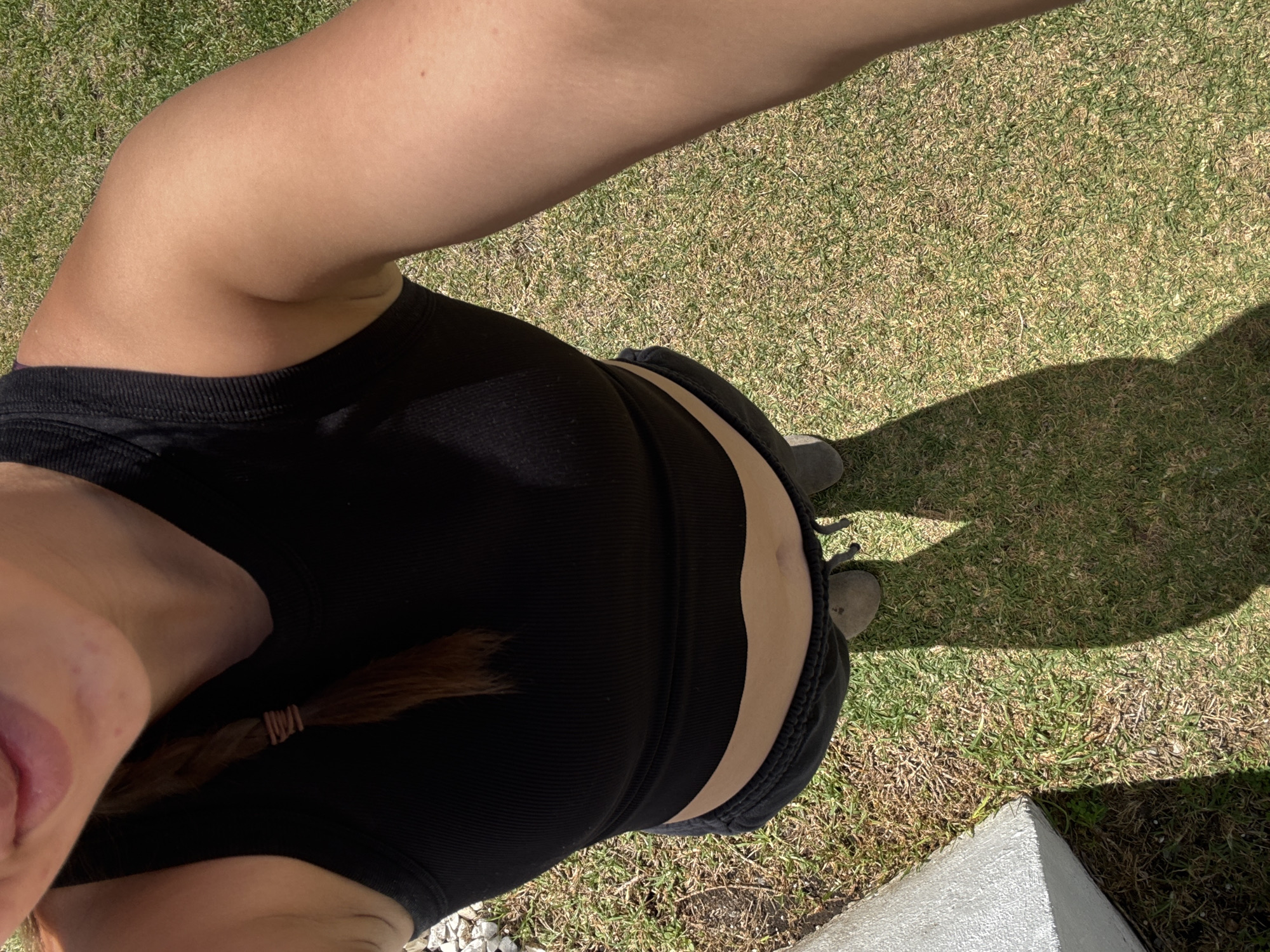 A first-person, downward-angled perspective shows a person standing on a grassy area. The person is wearing a black tank top and dark shorts. Part of the person's face is visible on the left side of the frame, along with a brown braid. The person's arm is extended upward, presumably holding the camera. The background consists of grass, a shadow cast by the person, and a corner of a concrete structure in the lower right corner. The location is determined to be Heroica Puebla de Zaragoza, Mexico, based on visual context clues.